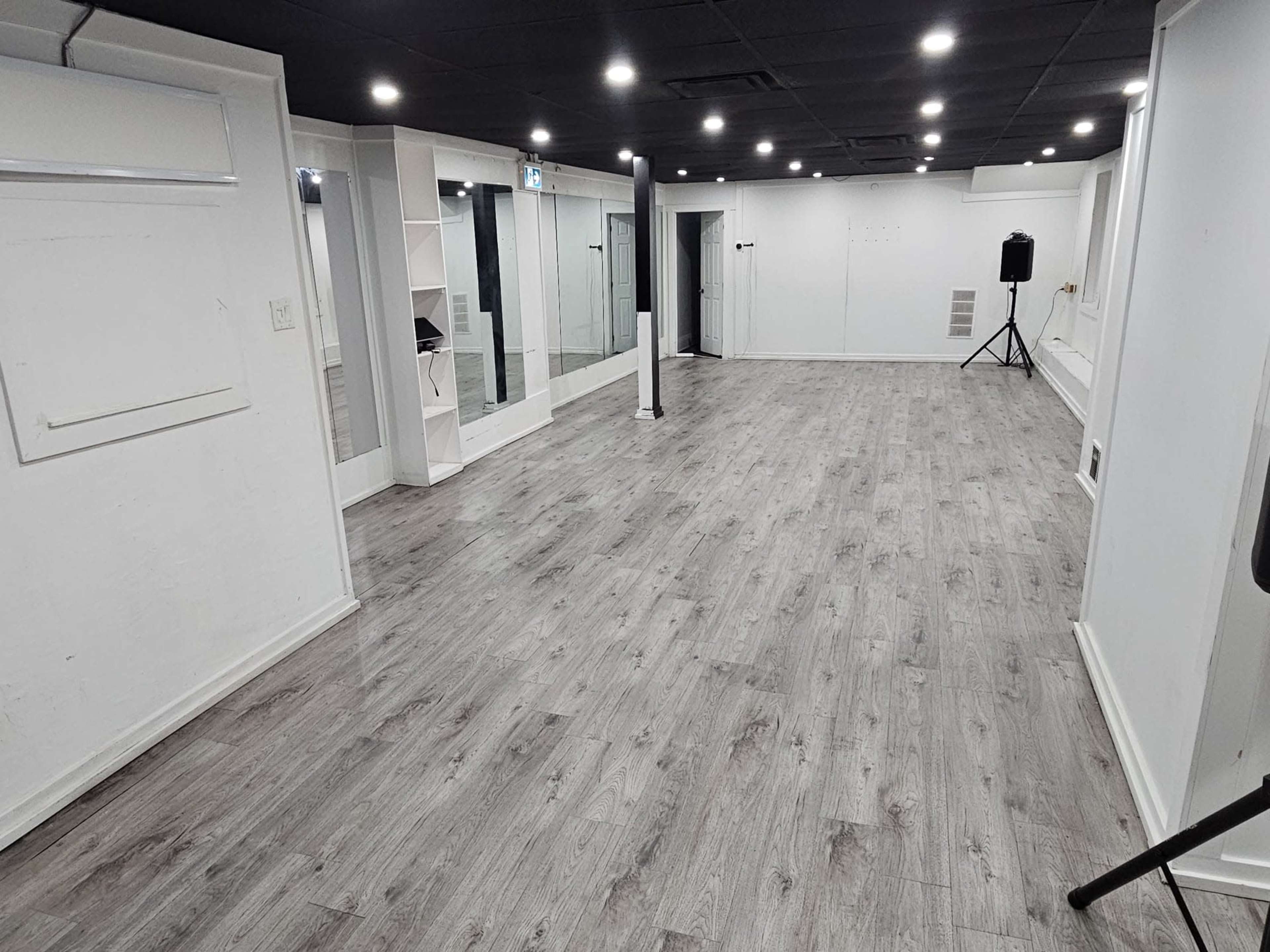 The image shows an empty room with wooden flooring, mirrors on one wall, and overhead lighting.