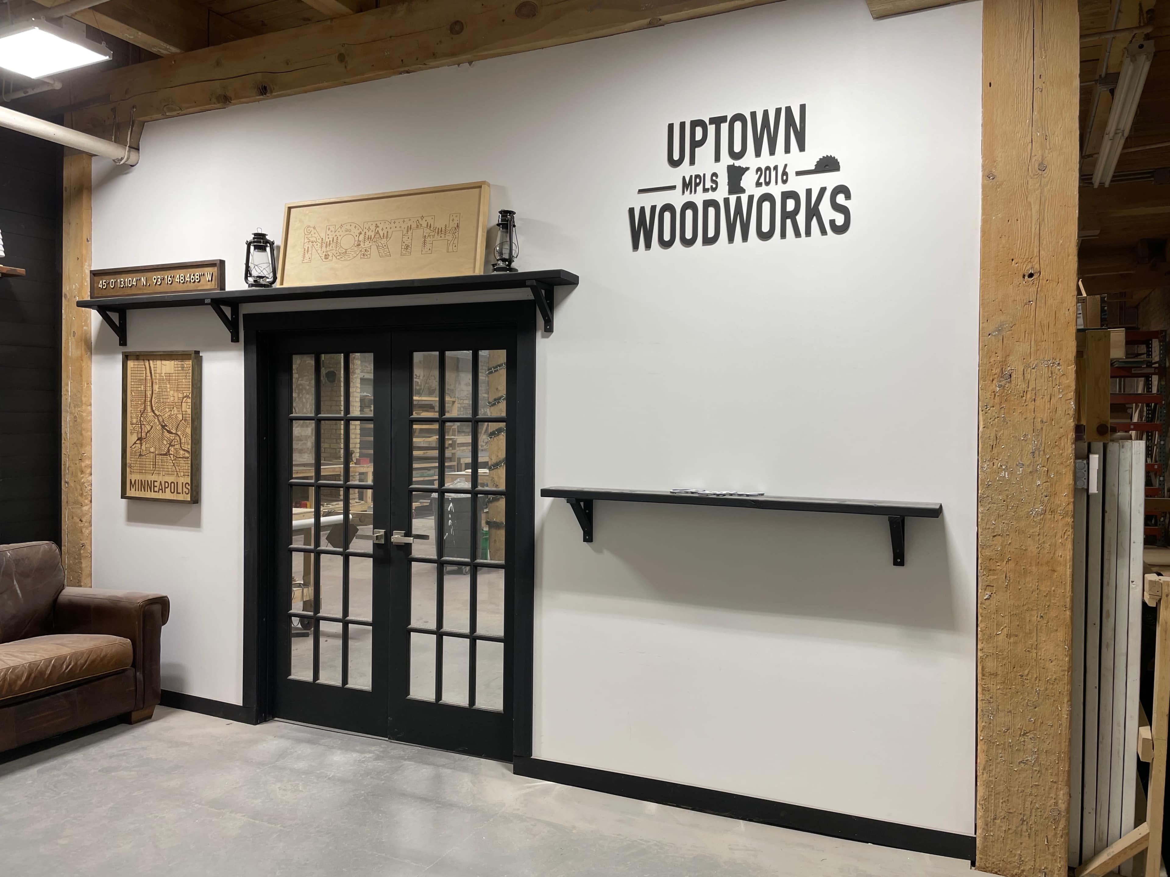 The image shows a wall featuring a sign that reads "UPTOWN WOODWORKS" with a framed map and a shelf underneath, alongside double glass doors in a wooden frame.