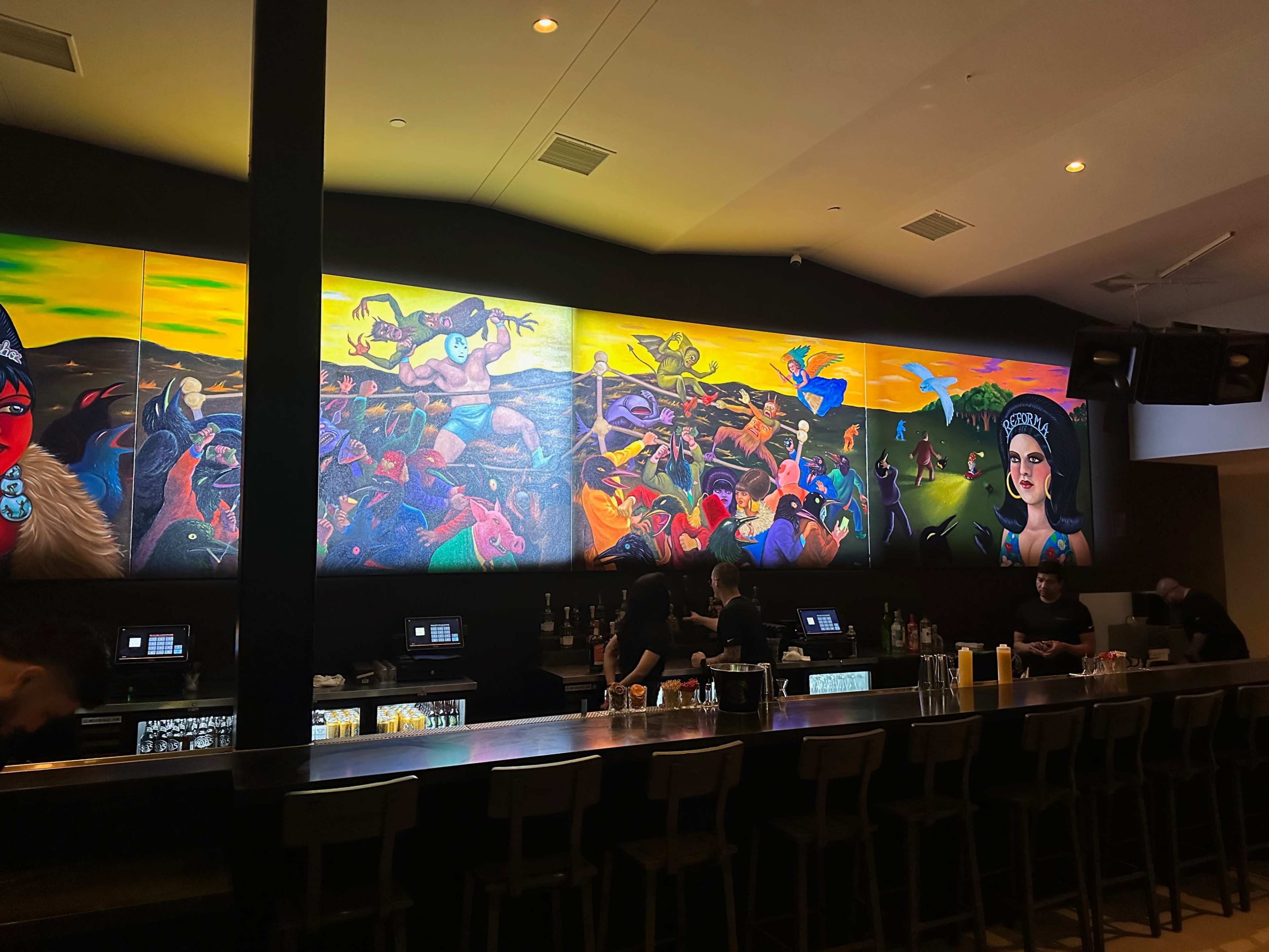 The image shows a vibrant mural on the wall of a bar, depicting various colorful figures and scenes, with bartenders working behind a long counter.