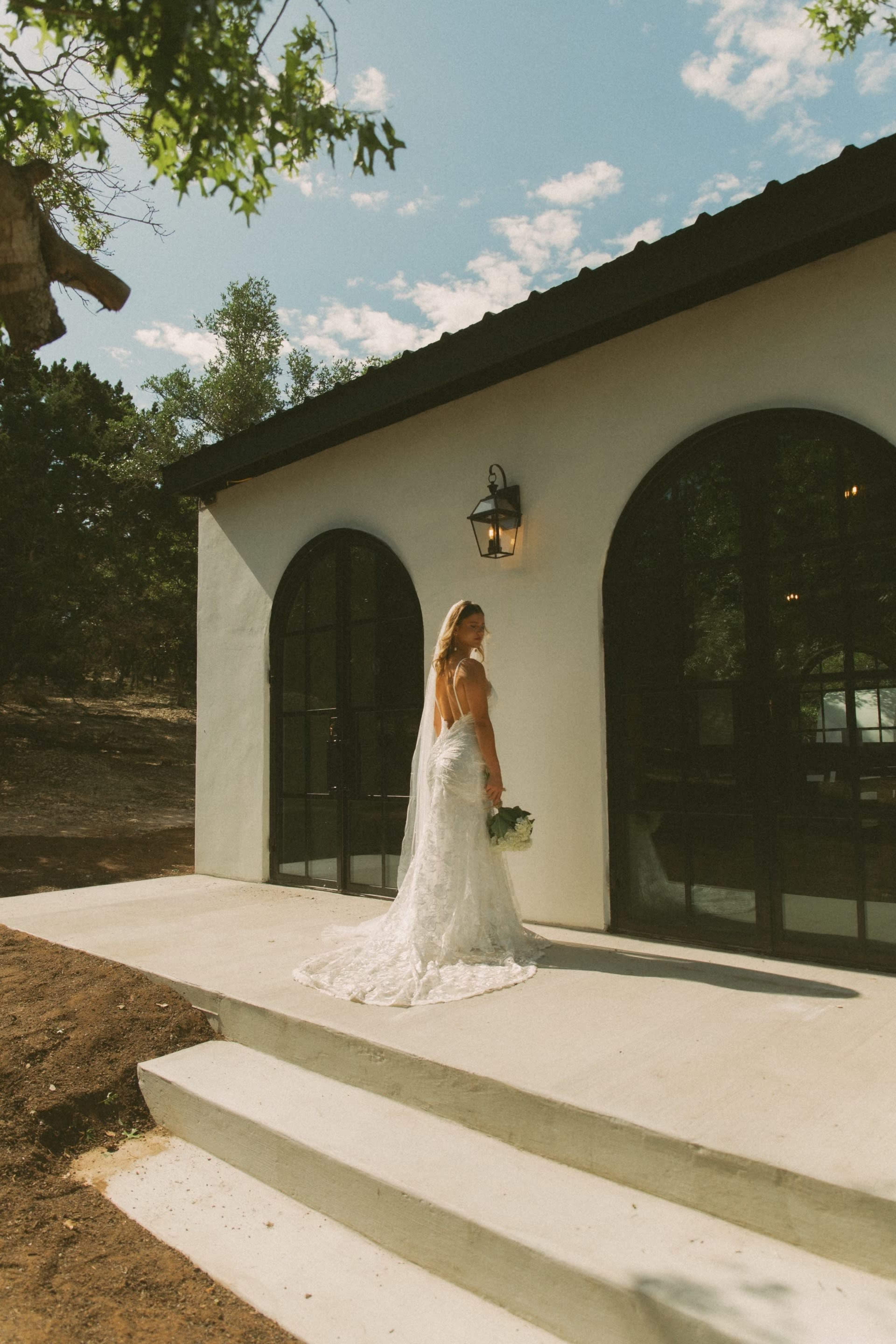A woman in a lace wedding dress stands on the steps of a white building with arched black windows, holding a bouquet of flowers.