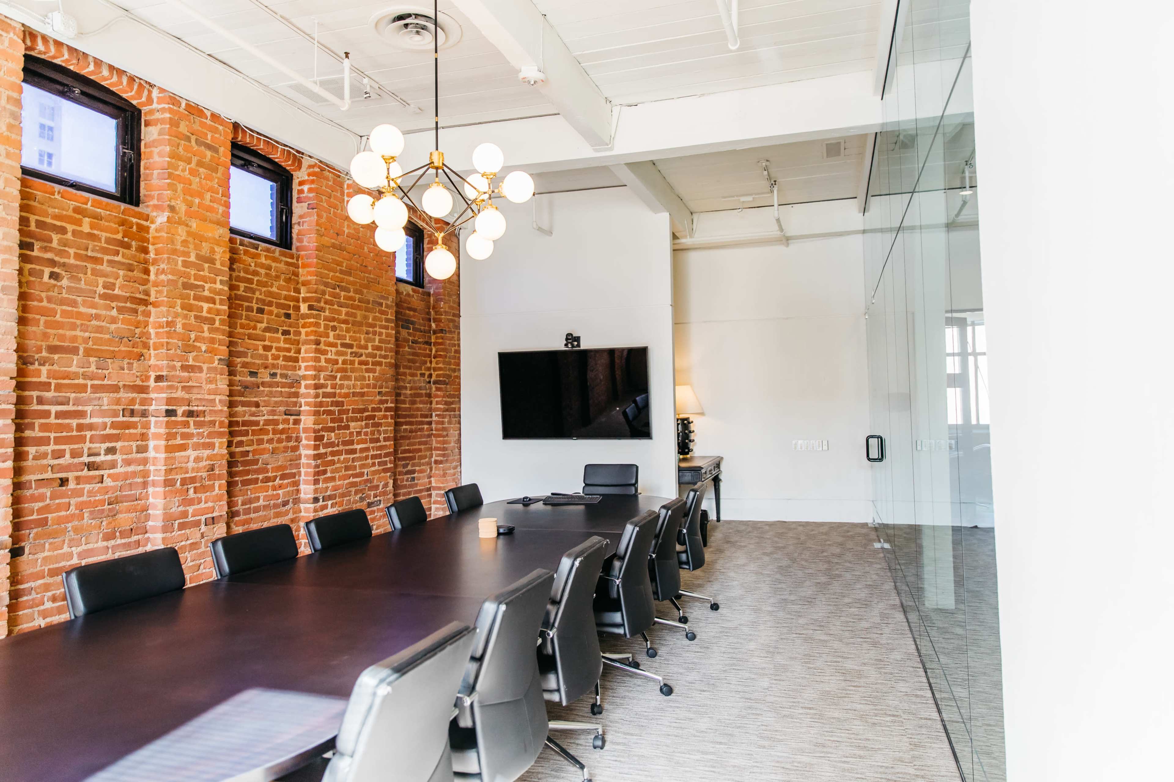 The image showcases a modern conference room featuring a long dark table surrounded by black chairs, brick walls, and a large screen mounted on the white wall.