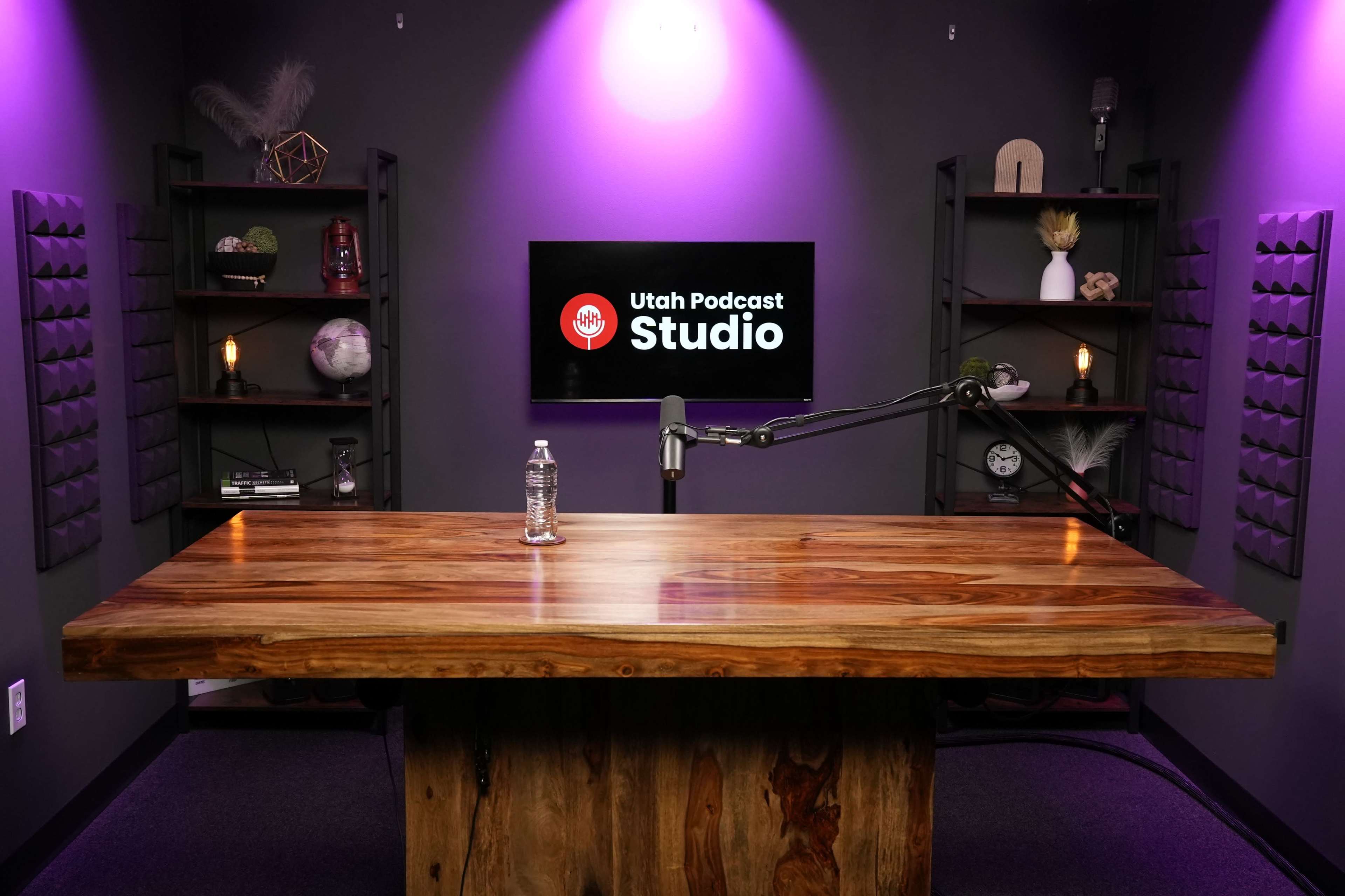 The image shows a podcast studio with a large wooden table, a microphone mounted on an arm, and a screen displaying "Utah Podcast Studio" against a backdrop of purple lighting and shelves containing various decorative items.