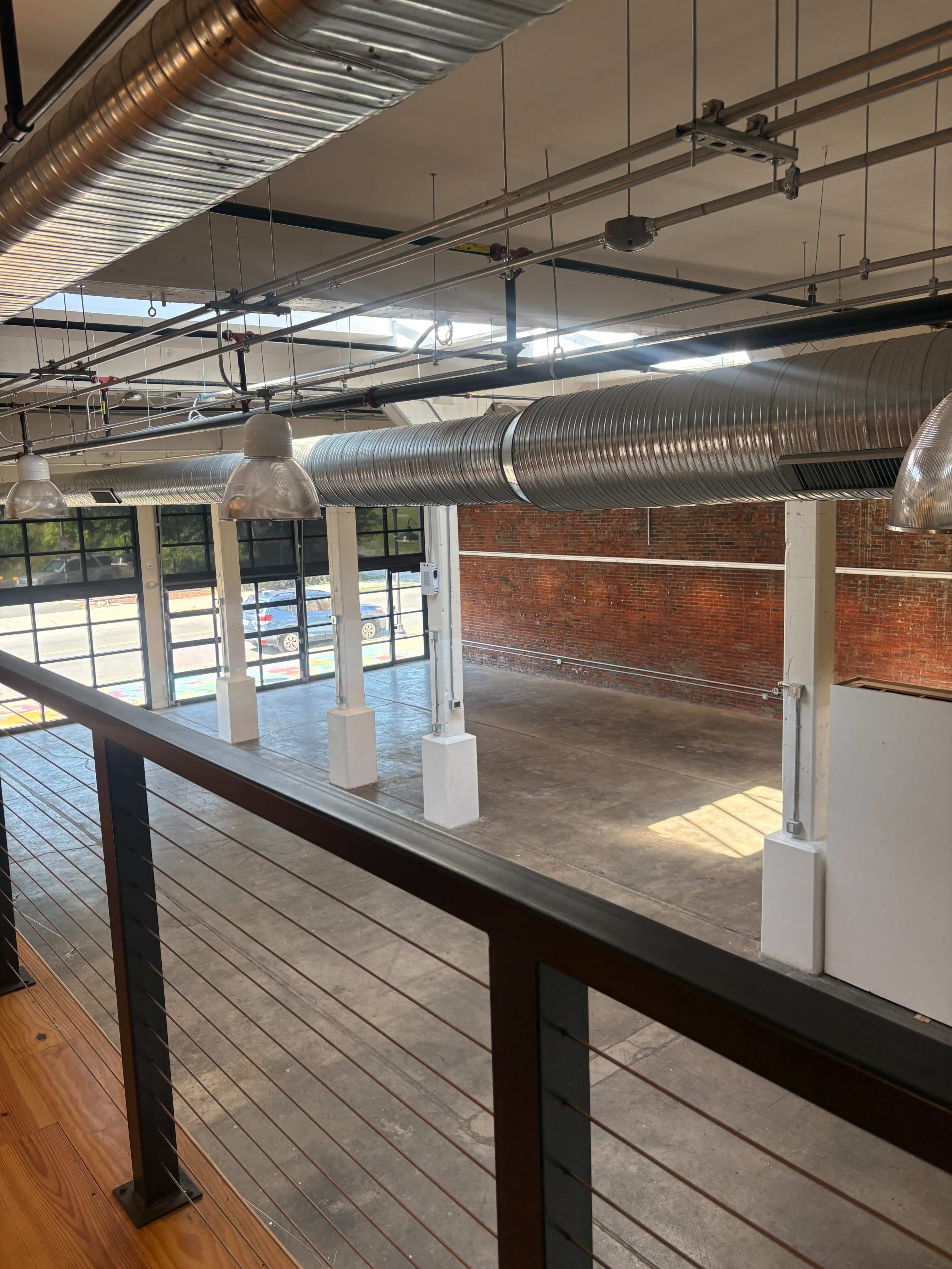The image shows an empty industrial space with exposed brick walls, large windows, and overhead metal ductwork.