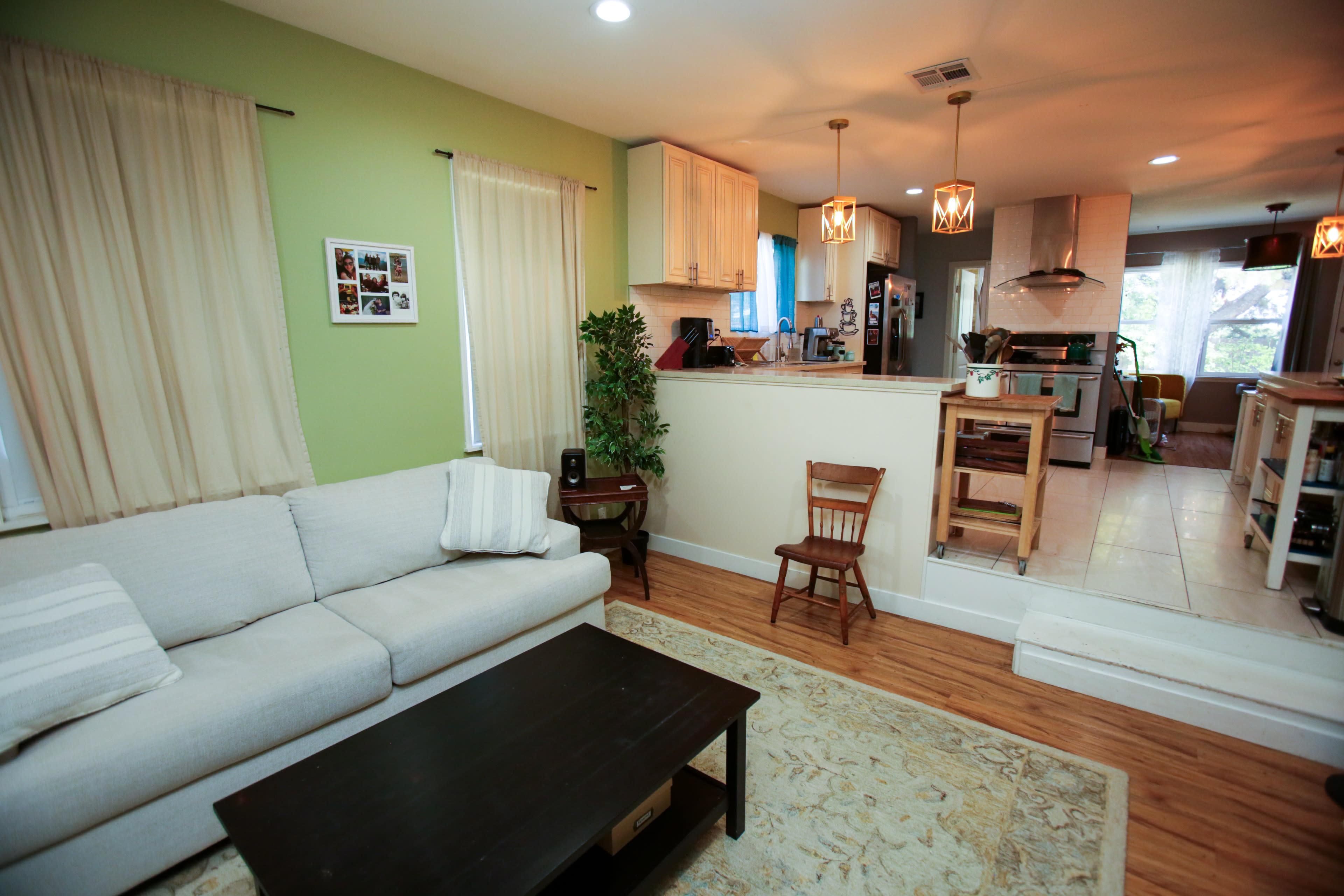 A cozy living room features a light-colored sofa, a dark coffee table, and a small wooden chair, with green and cream walls and an open kitchen area in the background.