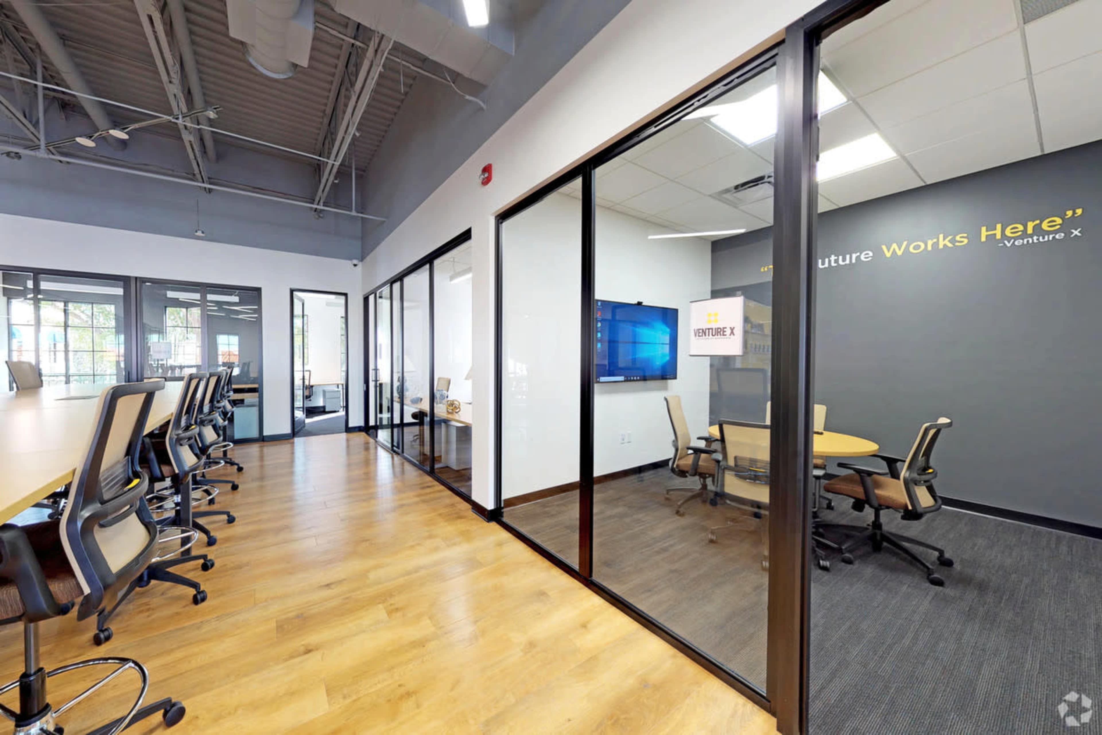 The image shows a modern office space featuring glass-walled meeting rooms and a collaborative area with conference tables and ergonomic chairs.