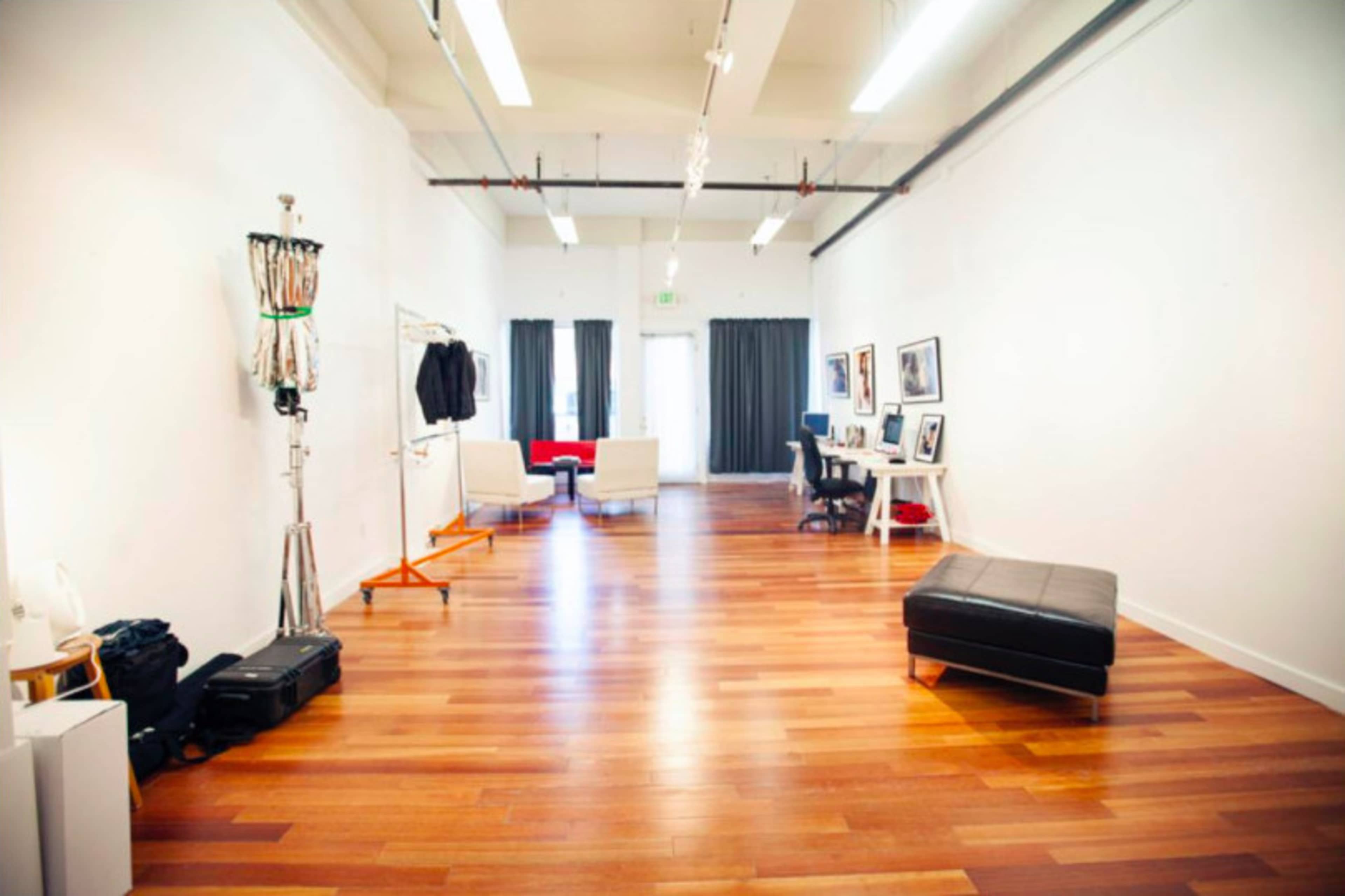 A spacious, well-lit room with wooden flooring, featuring a seating area, a desk setup, and a clothing rack.