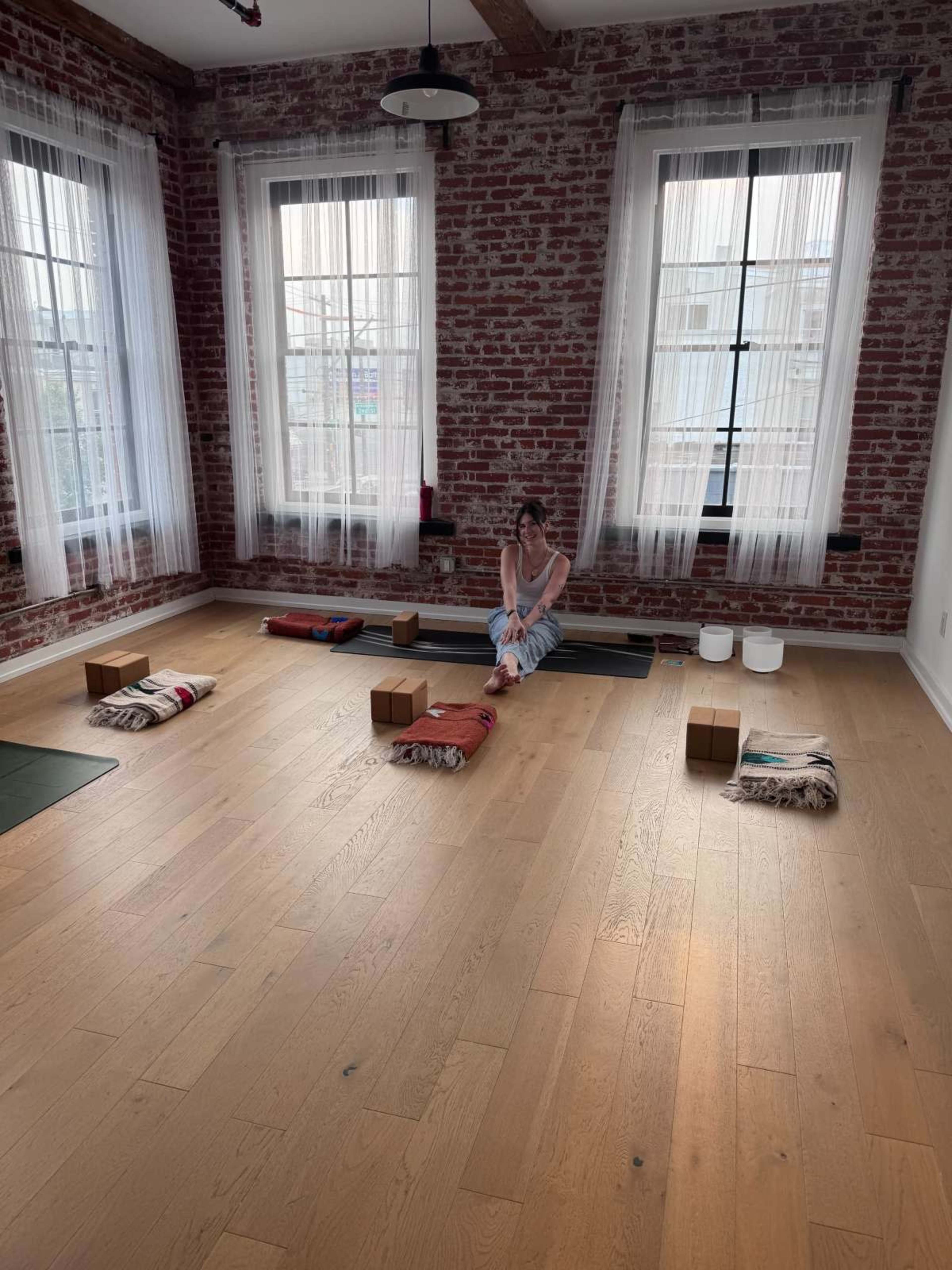A woman sits on the floor of a spacious room with exposed brick walls, surrounded by yoga mats and blocks.