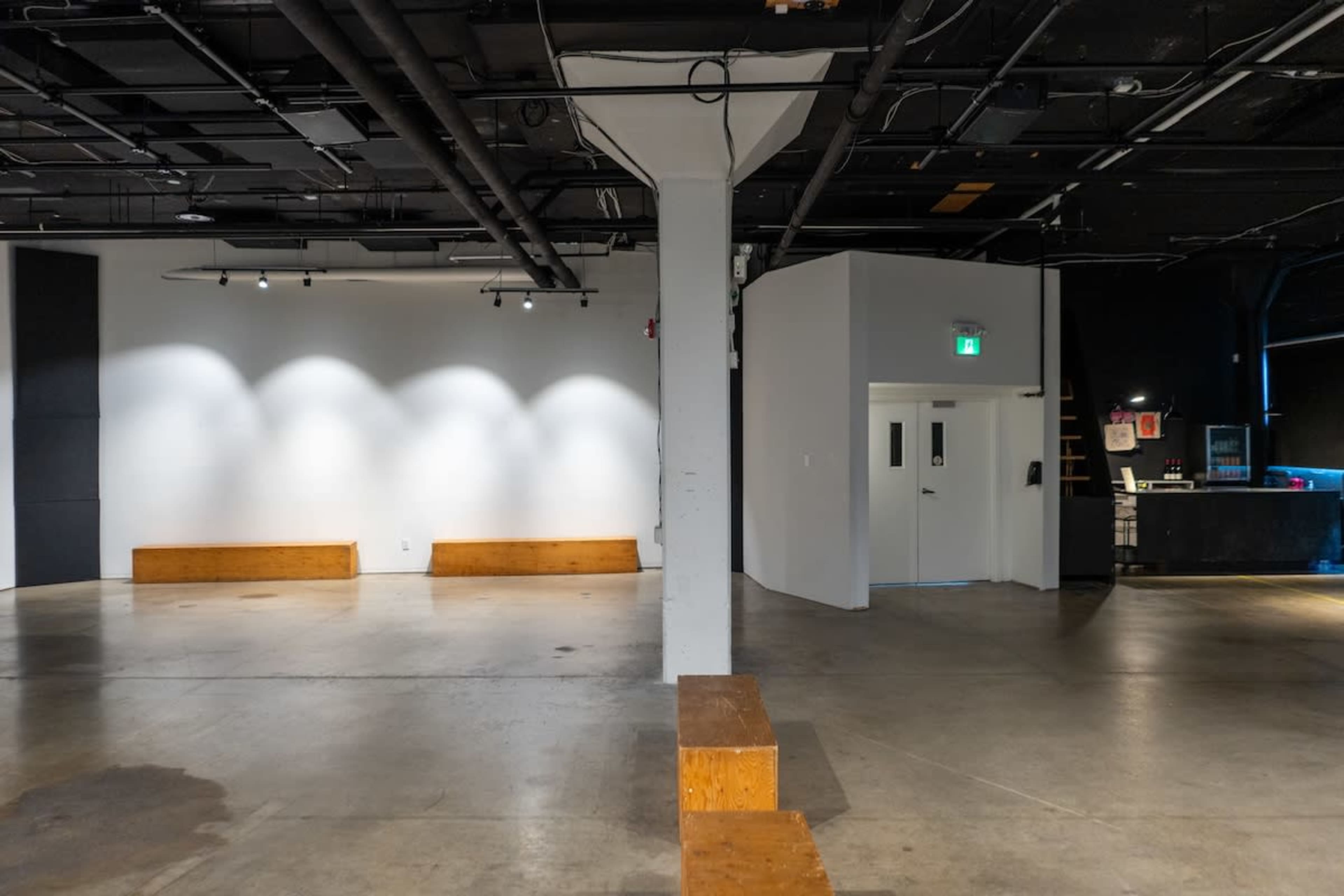The image shows an empty interior space with gray concrete flooring, a white wall with lighting, wooden benches, and a doorway leading to another area.