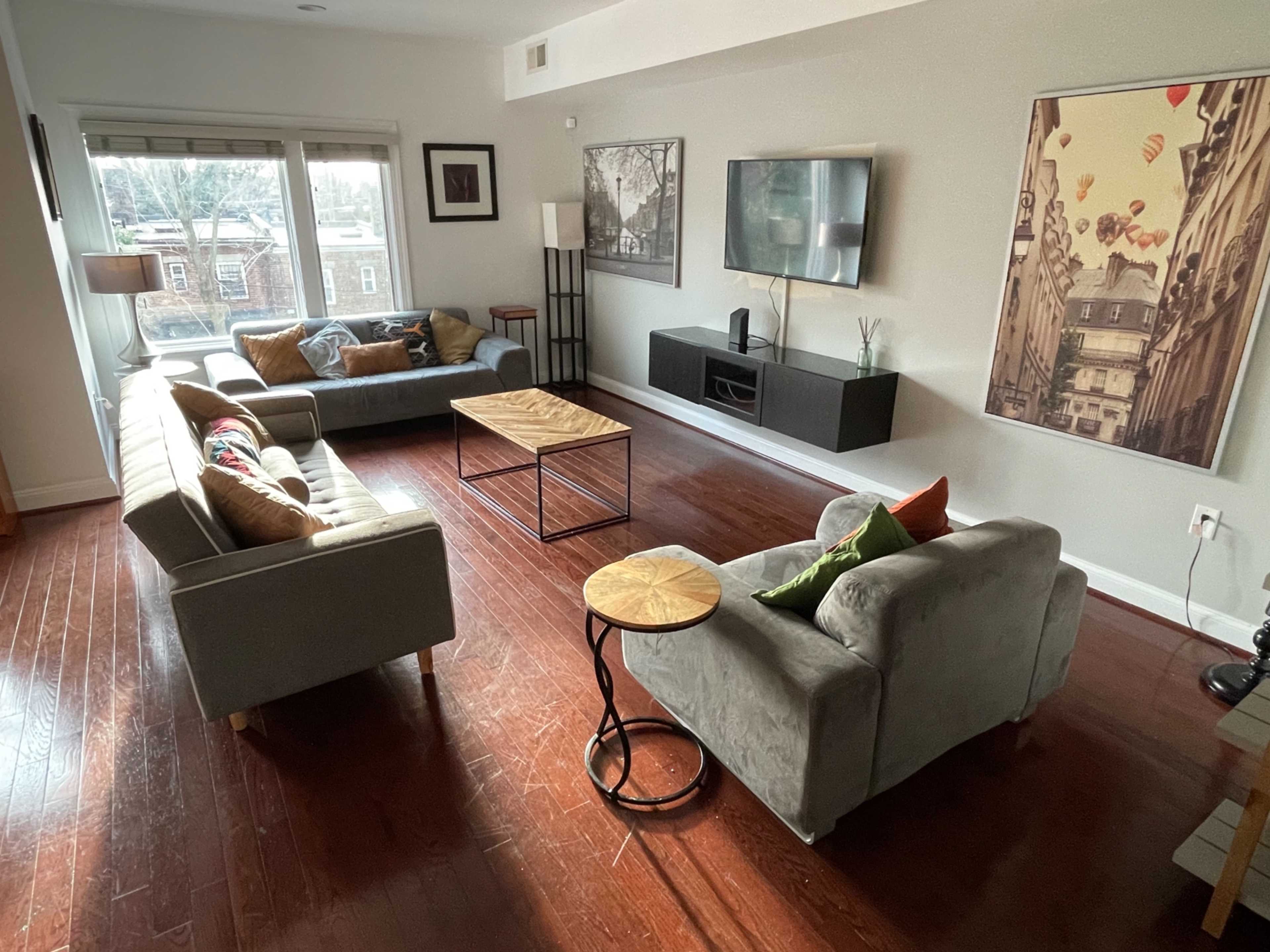 A modern living room with two sofas, a coffee table, and a wall-mounted television, illuminated by natural light from large windows.