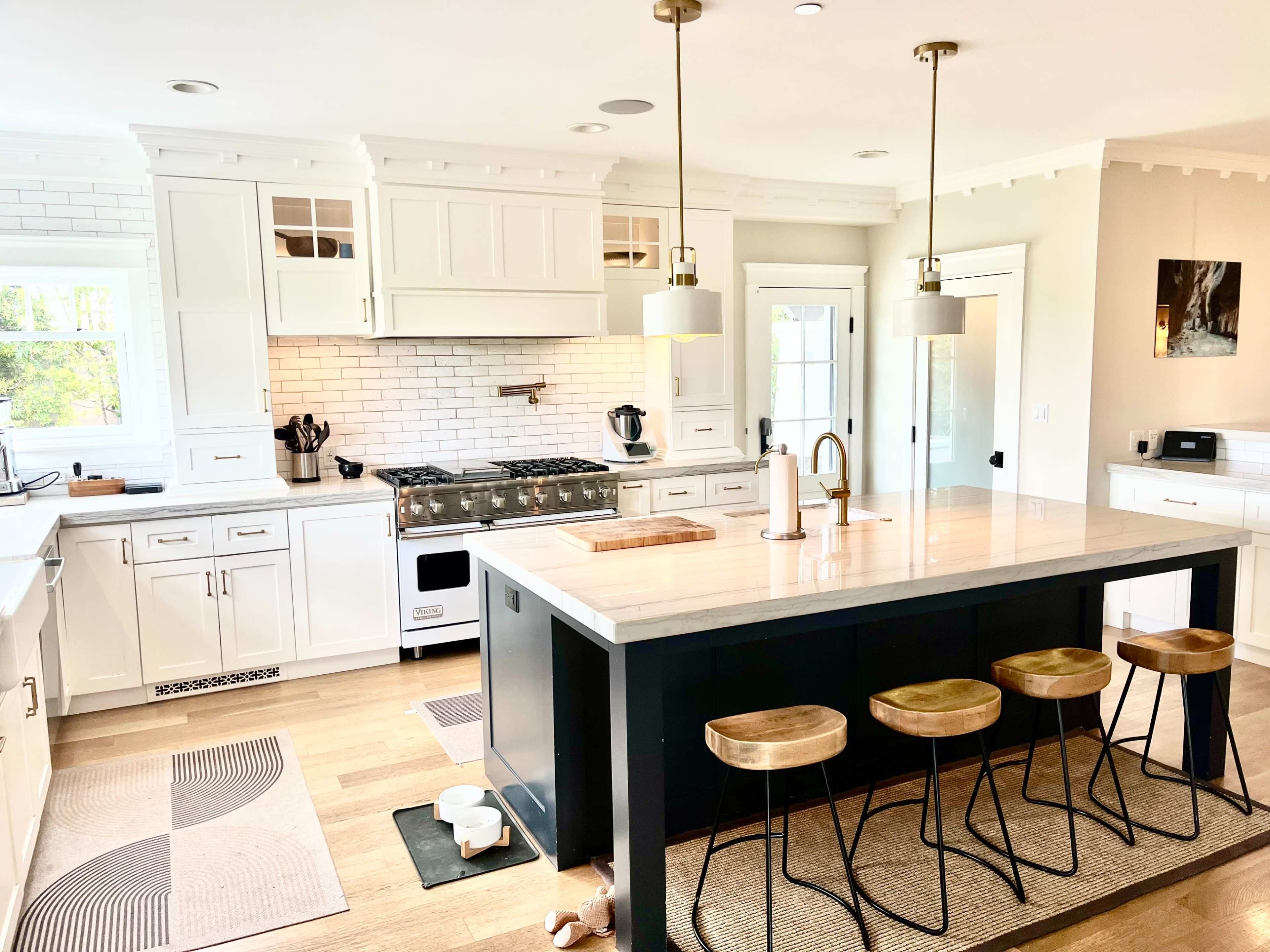 The image shows a modern kitchen featuring white cabinetry, a large island with wooden stools, and stainless steel appliances against a backdrop of light-colored walls and a tiled backsplash.
