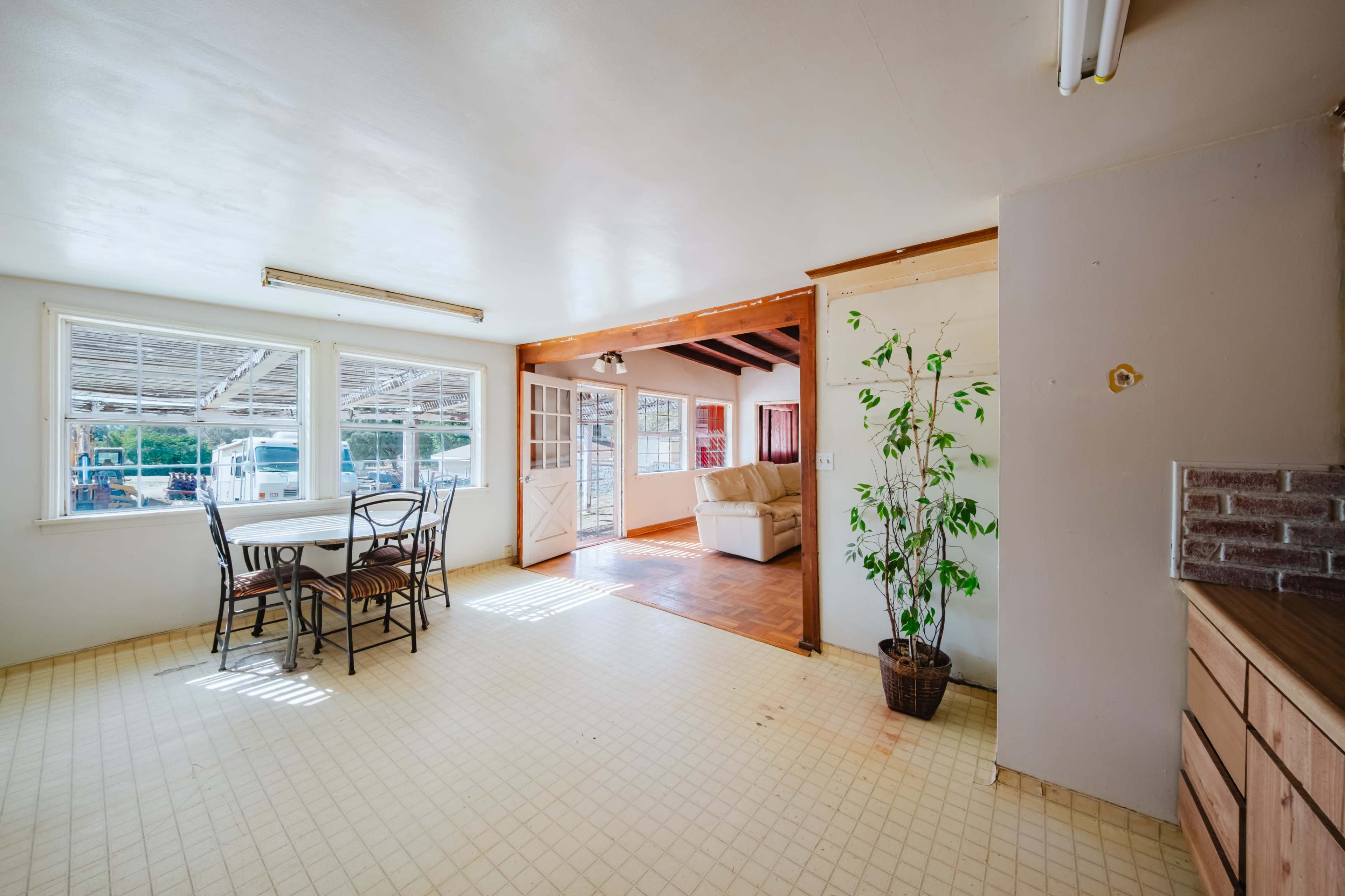 The image shows a spacious, well-lit room with a dining table and chairs, leading into another area that features a couch and large windows.