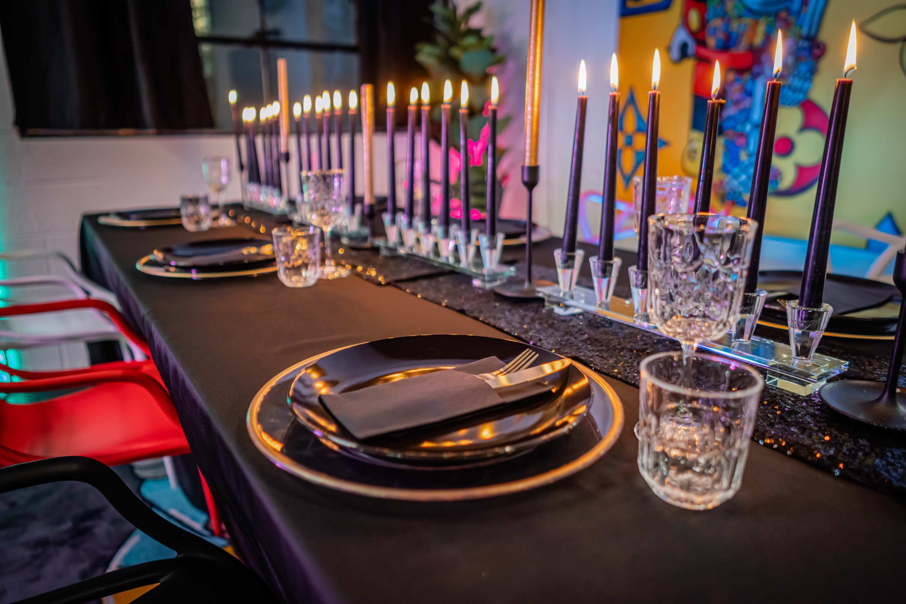 A long dining table is set with black plates, silverware, and clear glasses, illuminated by tall black candles placed along a shiny centerpiece.
