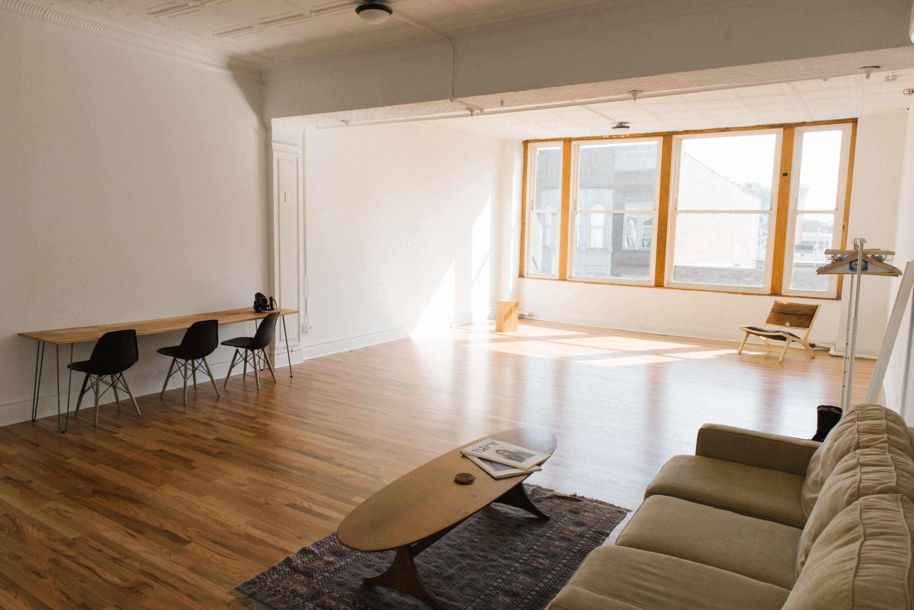 A spacious, minimalistic living room with a wooden floor, large windows providing natural light, a sofa, a coffee table, and a dining area with chairs.