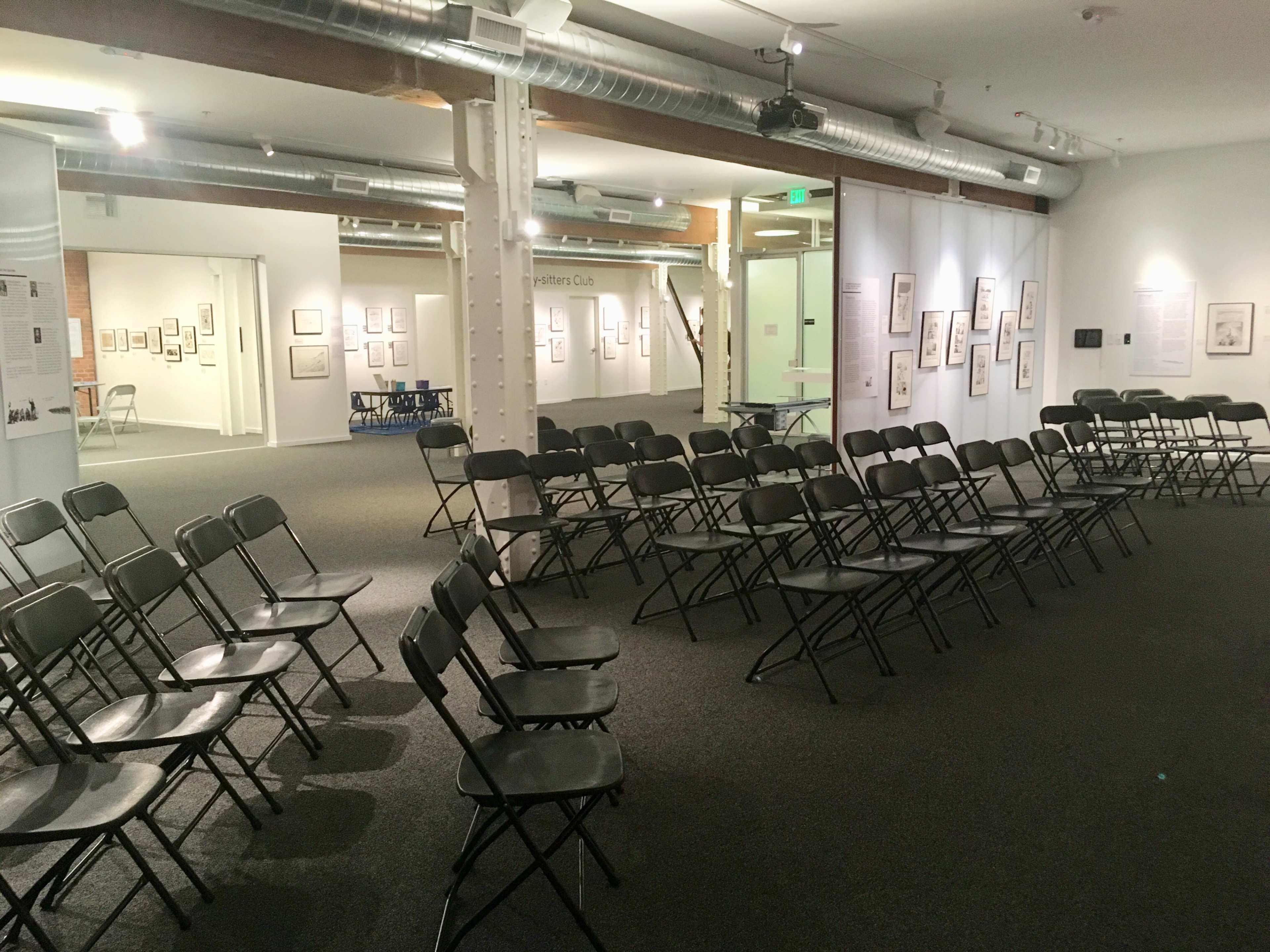 A series of black folding chairs are arranged in rows within a gallery space featuring framed artworks on the walls.