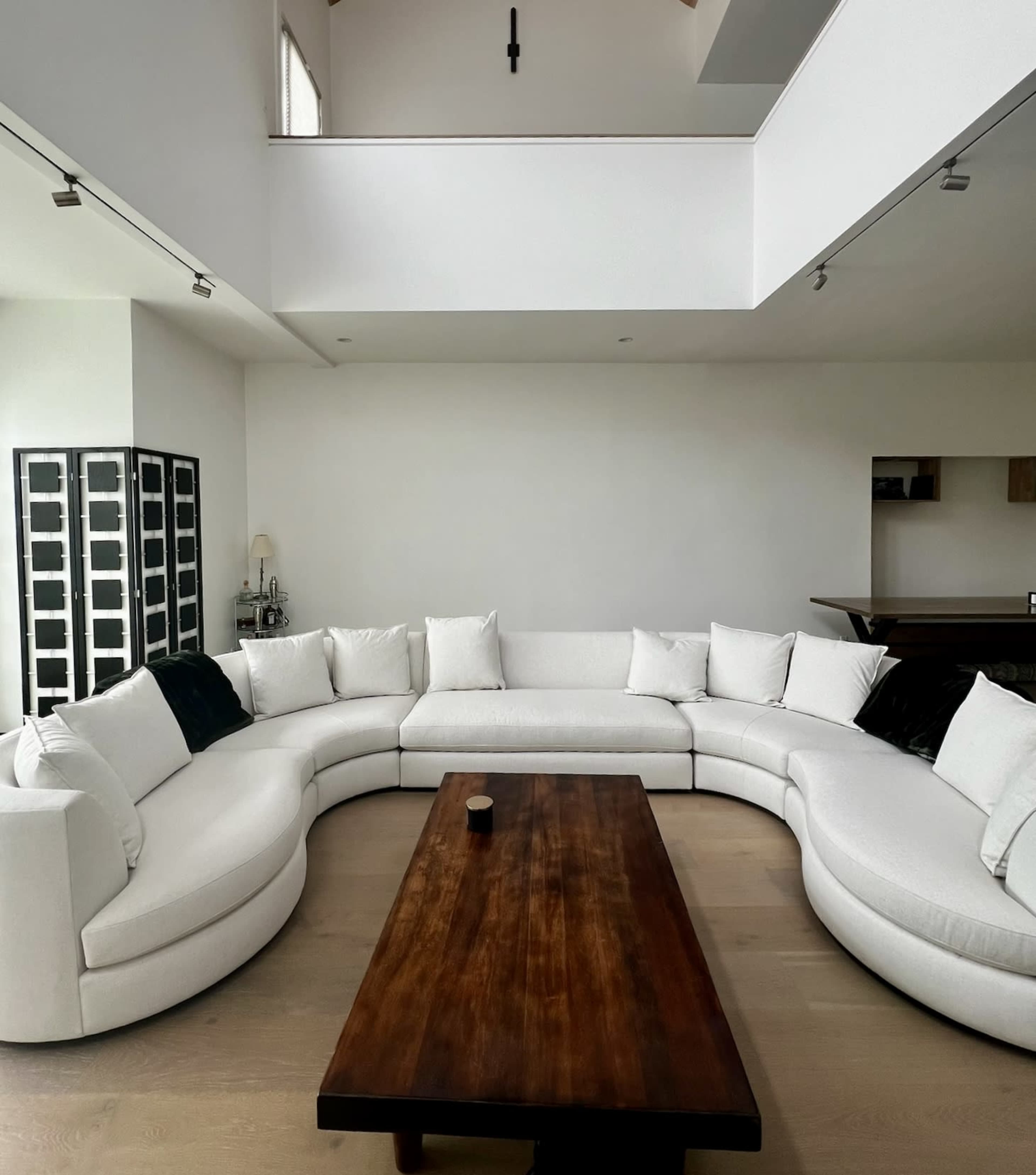 A modern, circular white sofa surrounds a dark wooden coffee table in a spacious, minimalistic living area with light hardwood flooring.