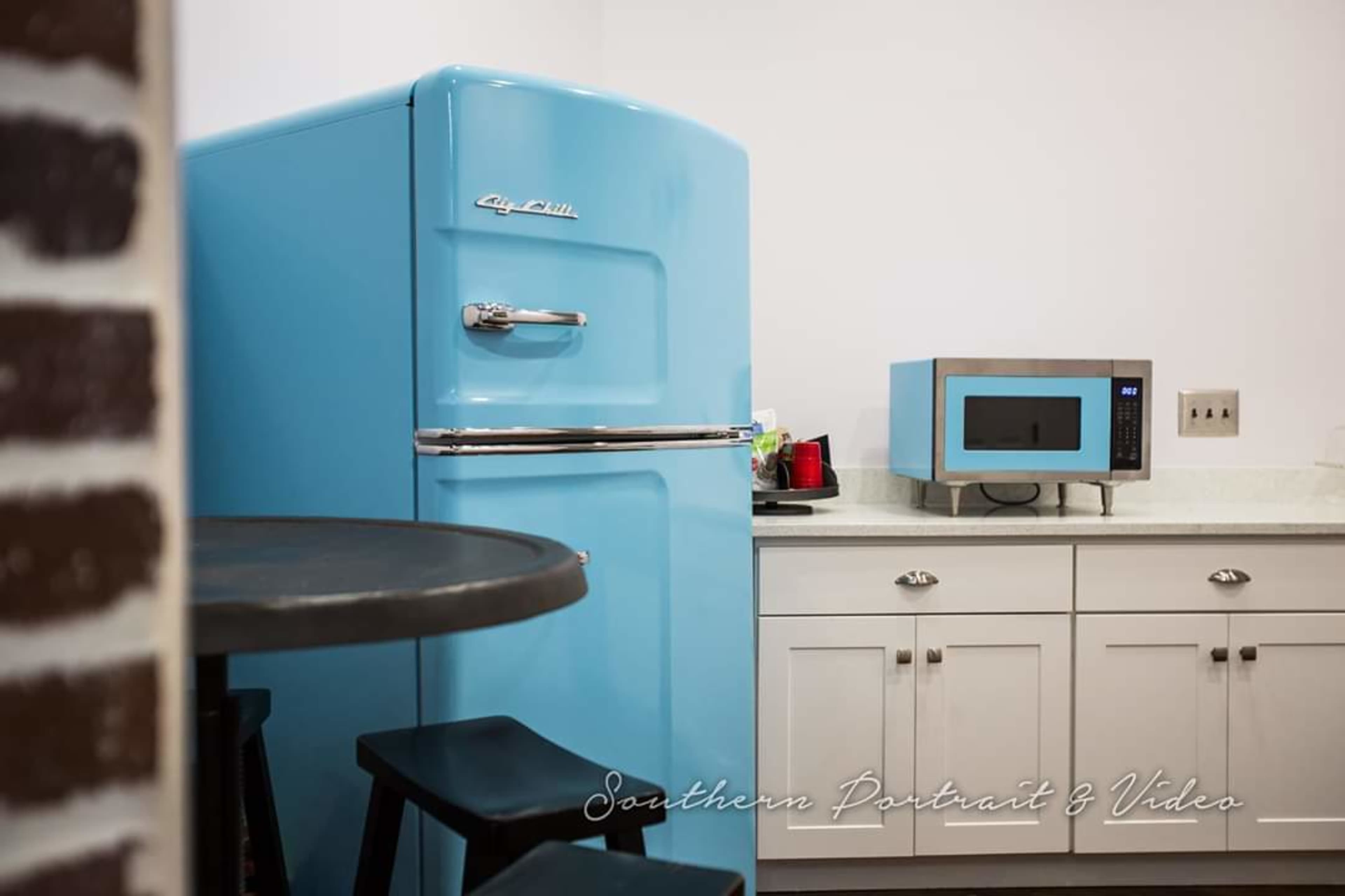 A bright blue vintage refrigerator stands next to a microwave on a countertop in a kitchen with white cabinetry and a dark table in the foreground.