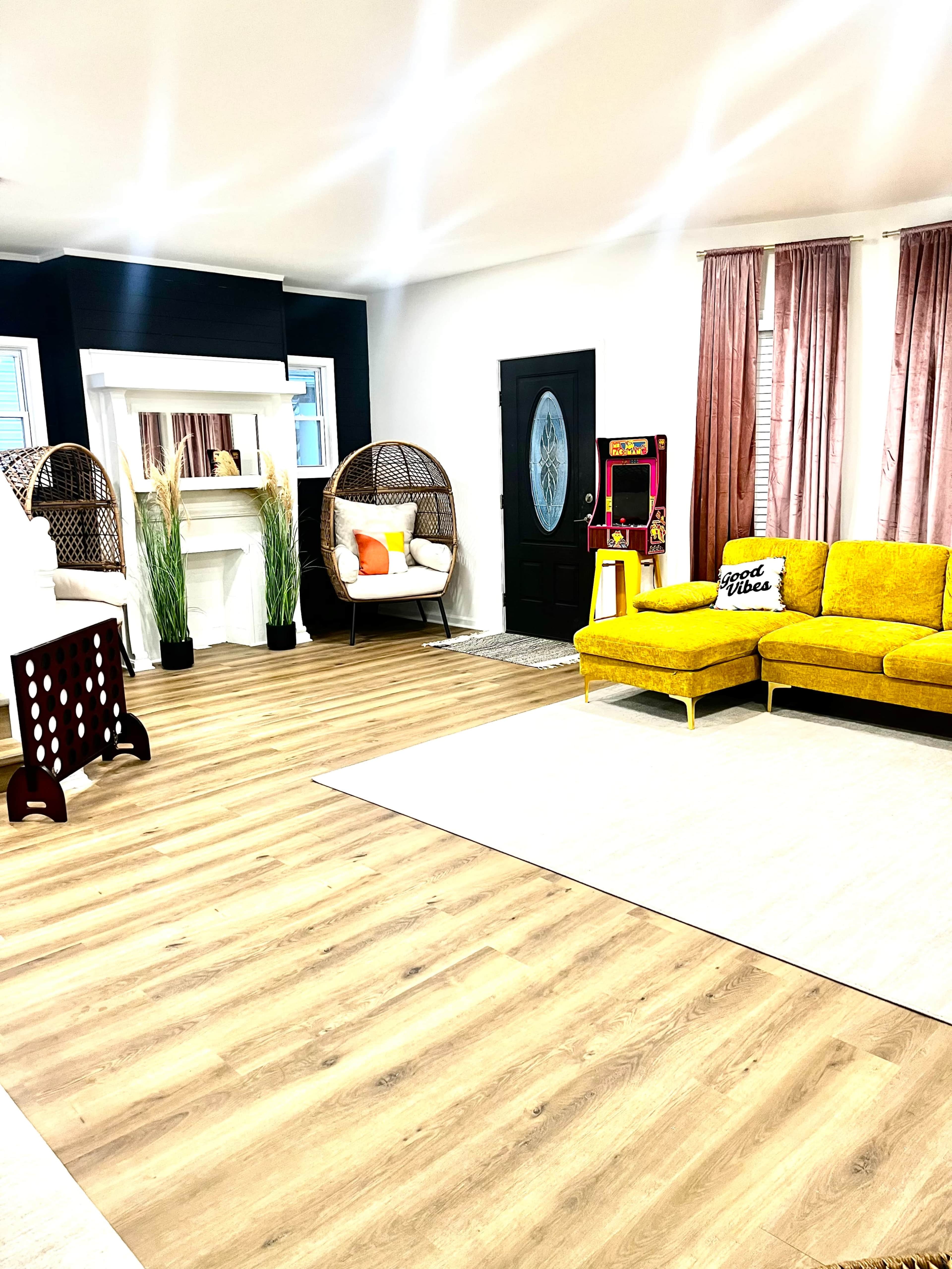 A spacious living room features a yellow couch, a white fireplace, wicker chairs, and a vintage arcade machine against a backdrop of light wood flooring.