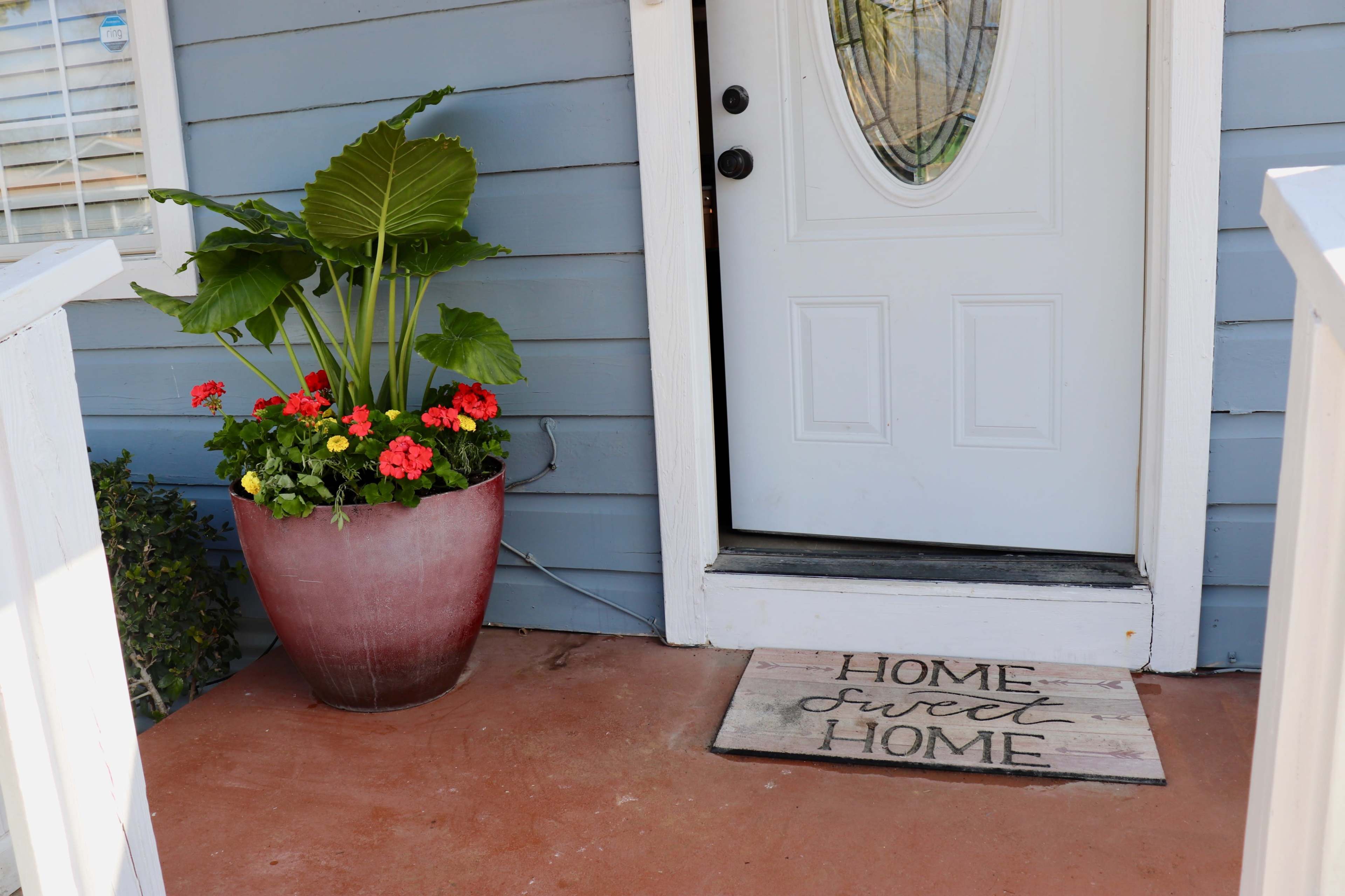 A front entryway features a potted plant with large leaves and red flowers beside a welcome mat that says "HOME sweet HOME."