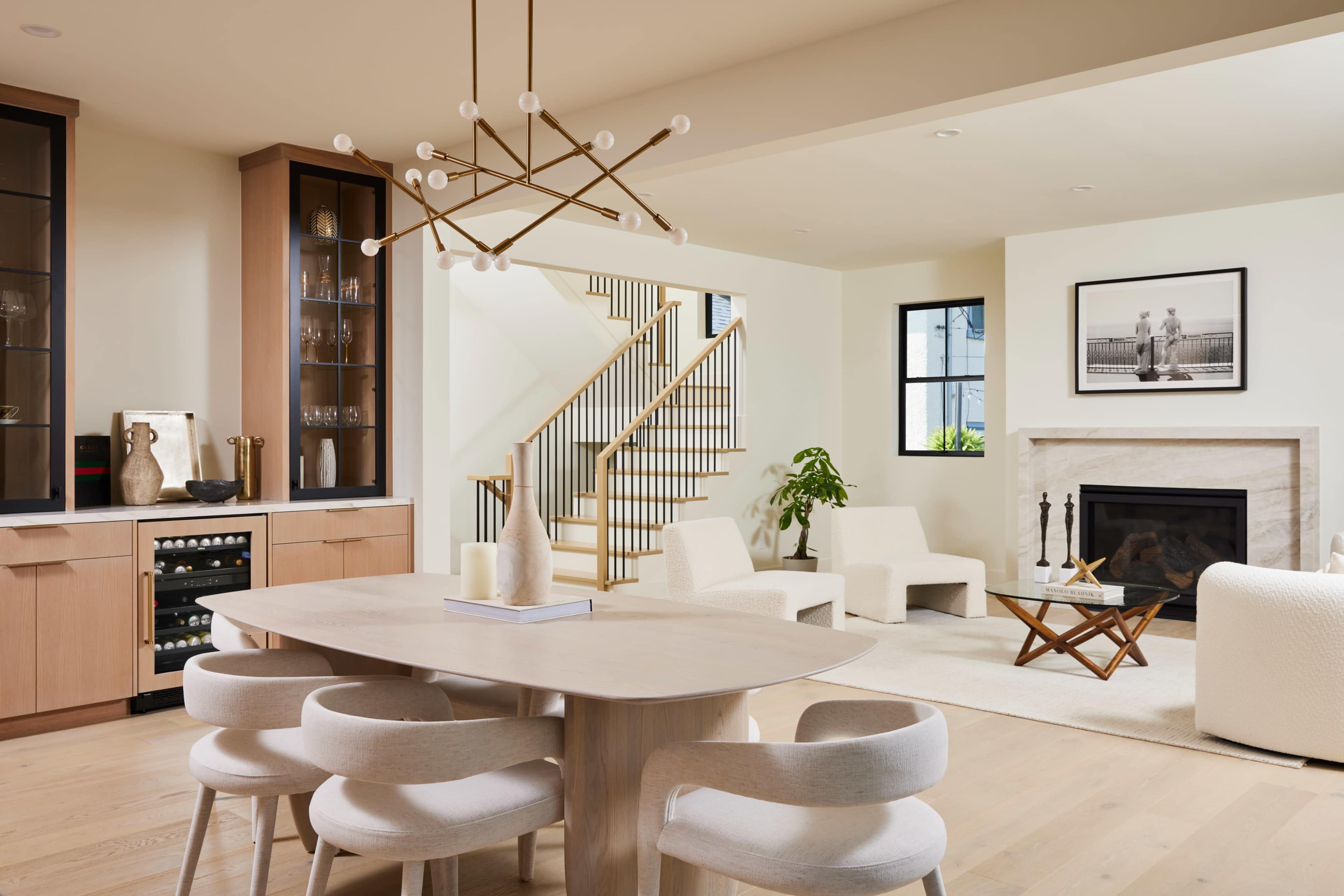 A modern dining area features a wooden table surrounded by curved chairs, with a staircase, cozy seating area, and a fireplace in the background.