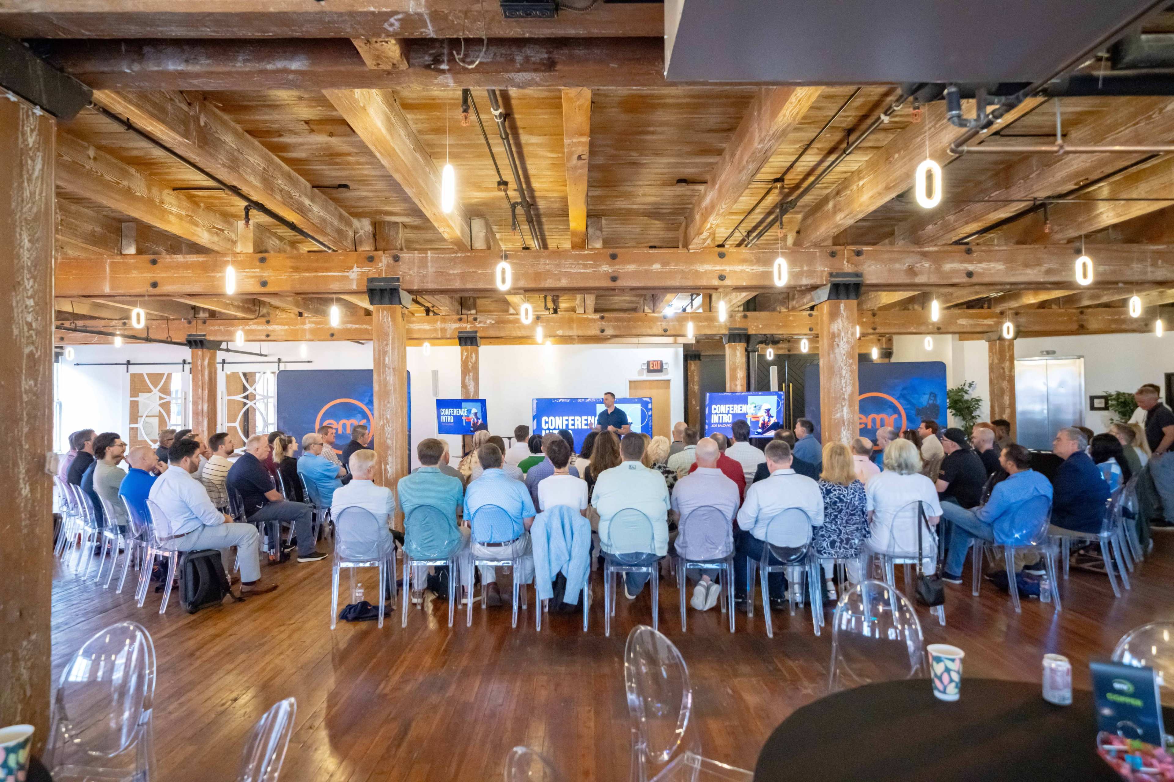 A large group of attendees is seated in a workshop or conference setting, facing a speaker at the front of a room with exposed wooden beams and modern decor.