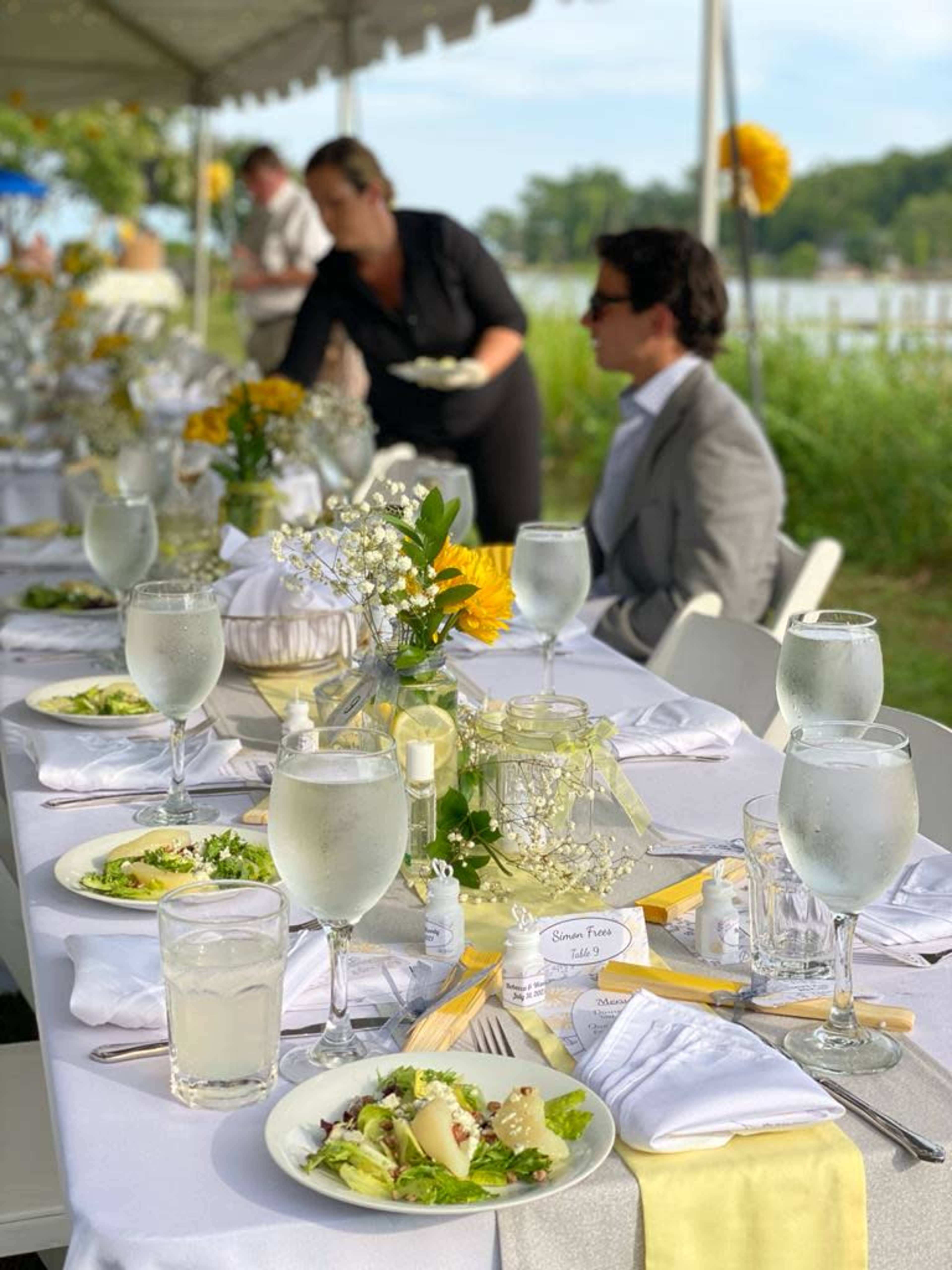 A long table elegantly set with glassware, floral decorations, and plates of salad occupies an outdoor space near a body of water, with a server in the background.