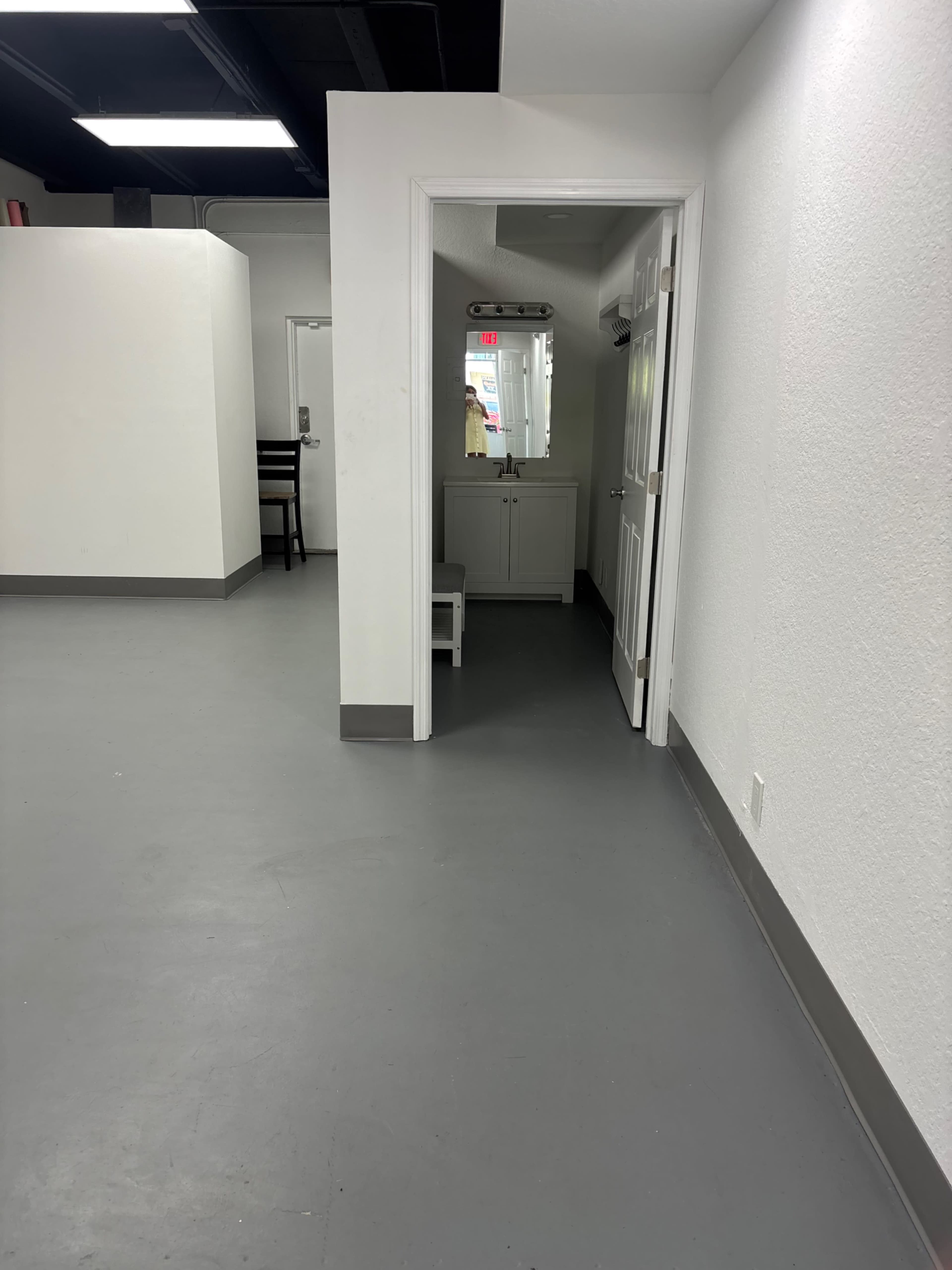 The image shows a minimalist hallway leading to an area with a mirror and a cabinet, flanked by white walls and gray flooring.