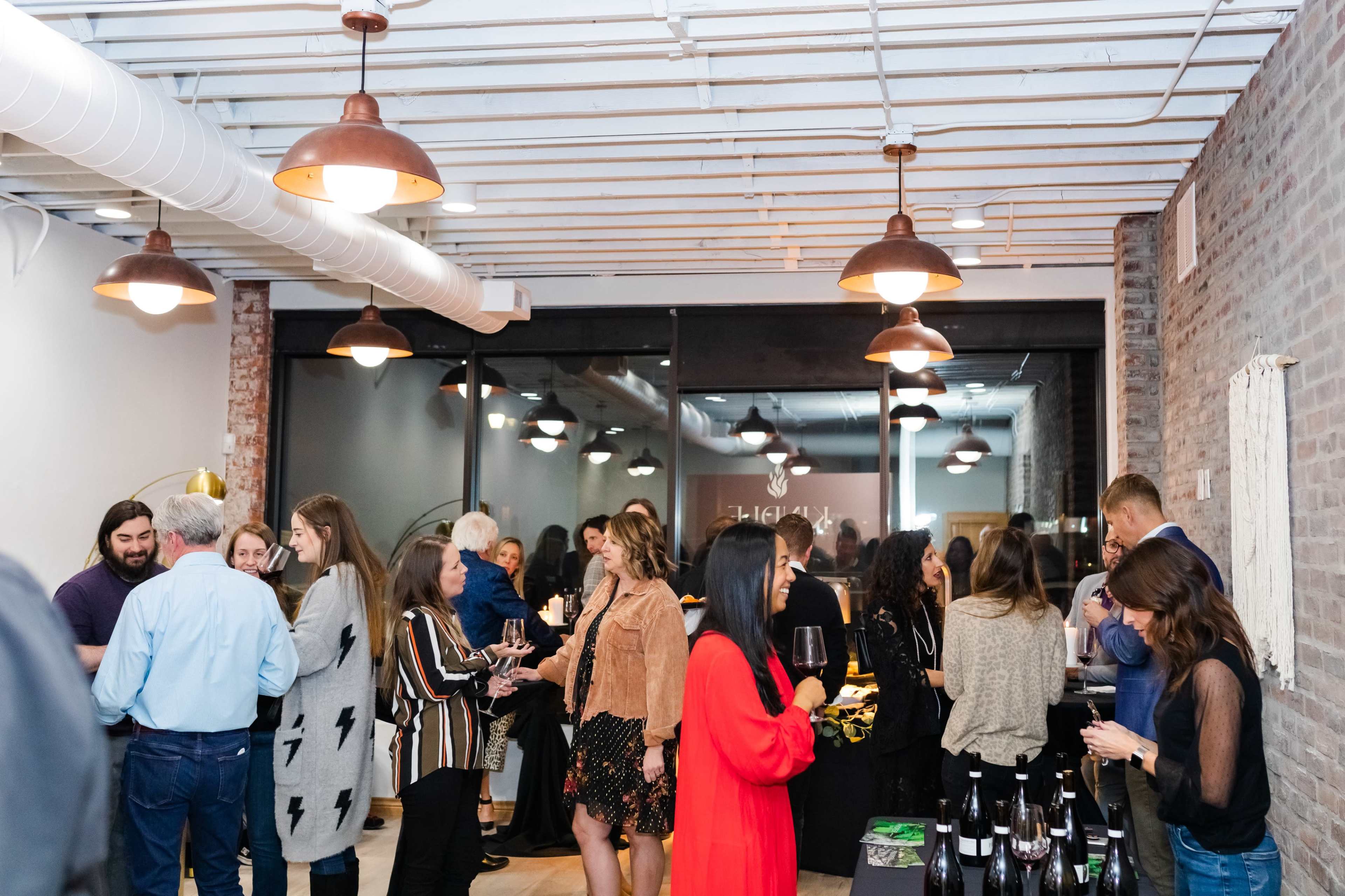 A group of people engages in conversation and socializing at a gathering in a modern space with exposed brick walls and pendant lights.