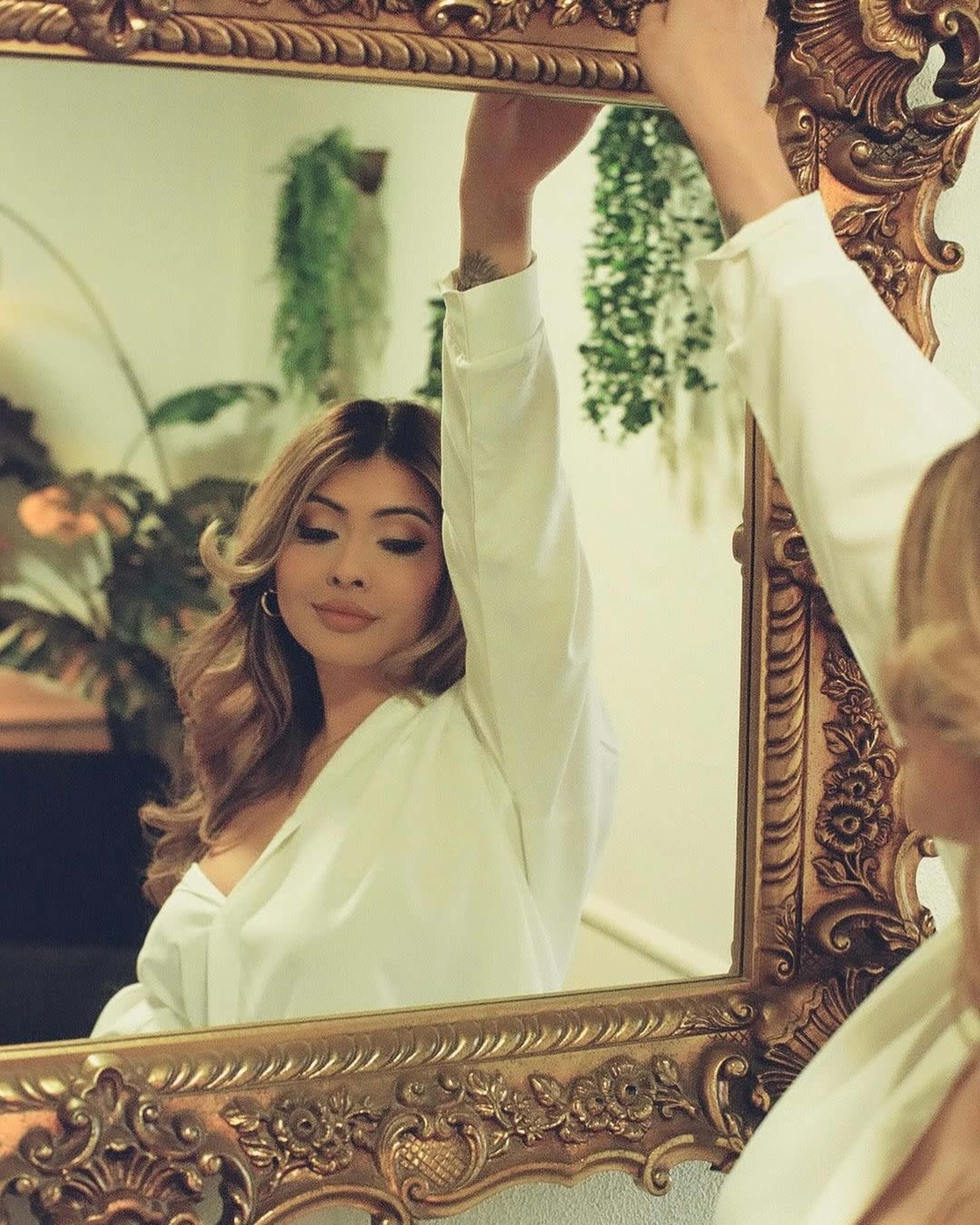 A person in a white shirt poses in front of an ornate mirror surrounded by greenery.
