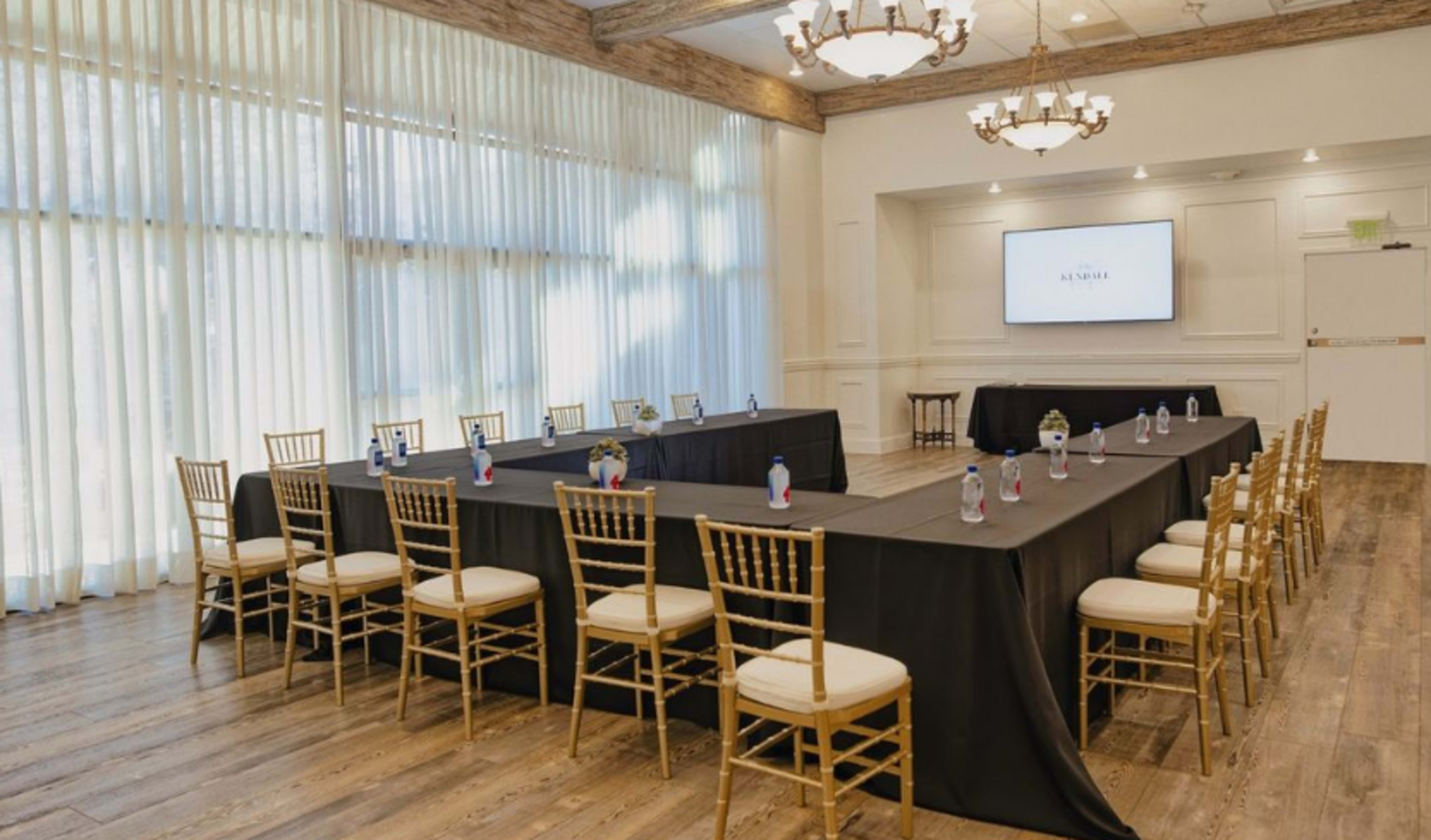 A conference room is set up with a large black table surrounded by gold chairs, featuring water bottles and a screen on the wall.