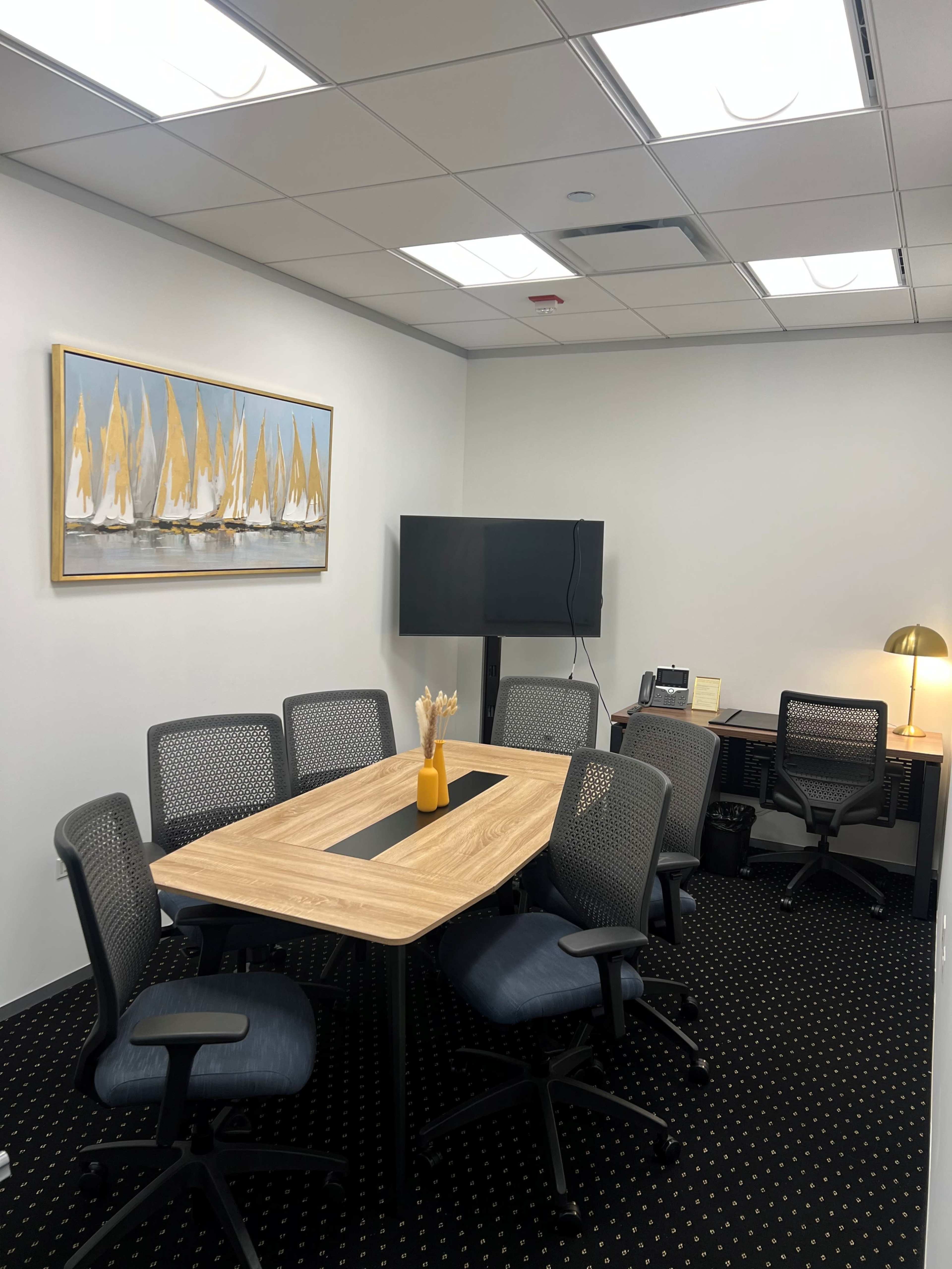 The image shows a small conference room featuring a wooden table surrounded by six mesh office chairs, a television mounted on the wall, and a decorative painting of sailboats.