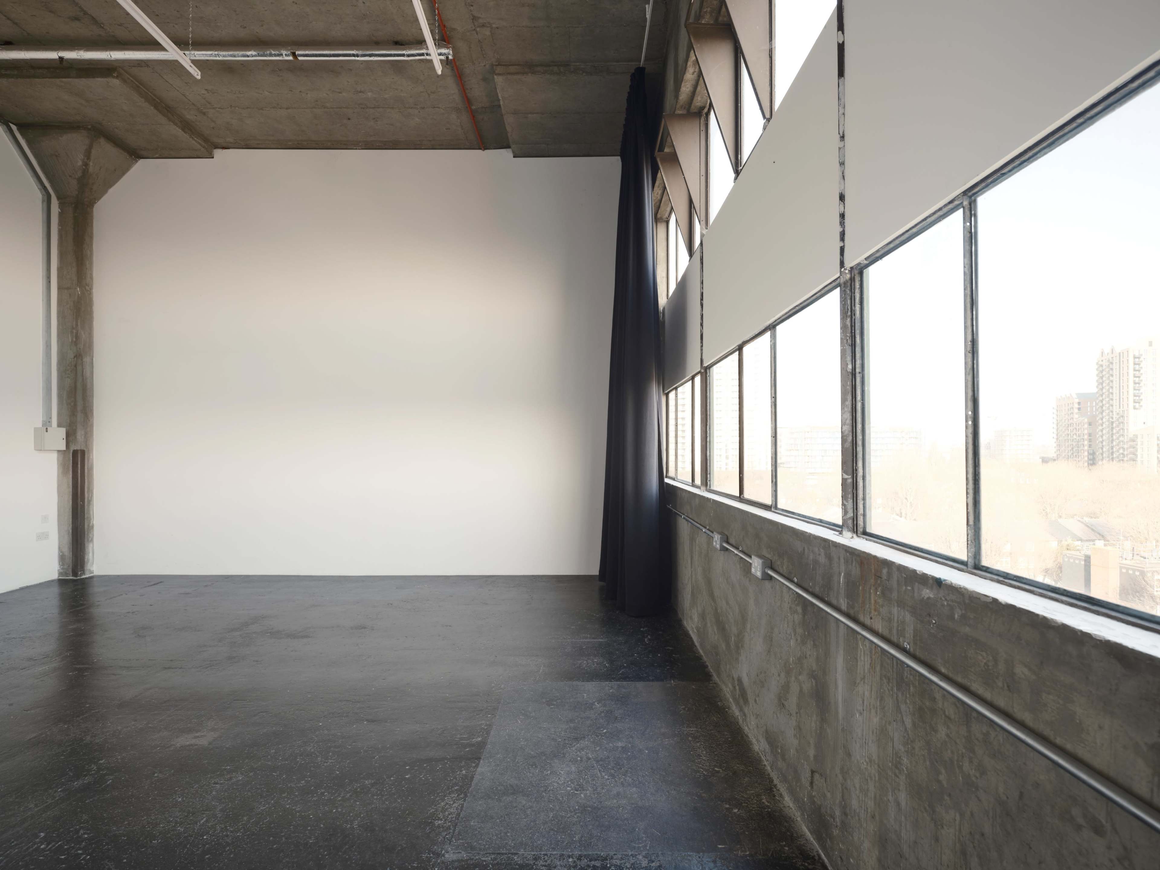 A spacious, industrial-style room with large windows, concrete walls, and a dark curtain partially drawn.