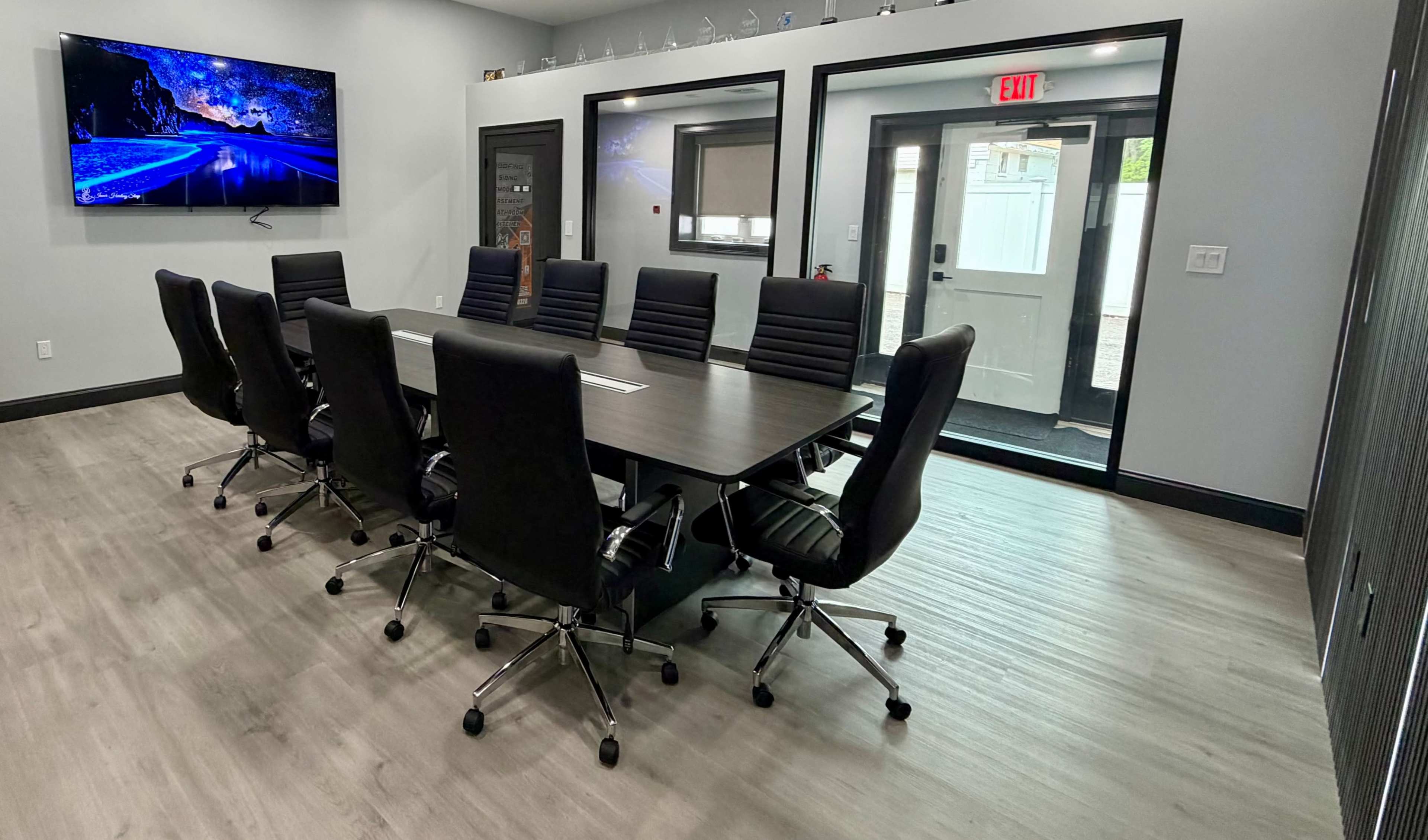 A modern conference room features a long table surrounded by black ergonomic chairs, with a large television mounted on the wall and a door leading outside.