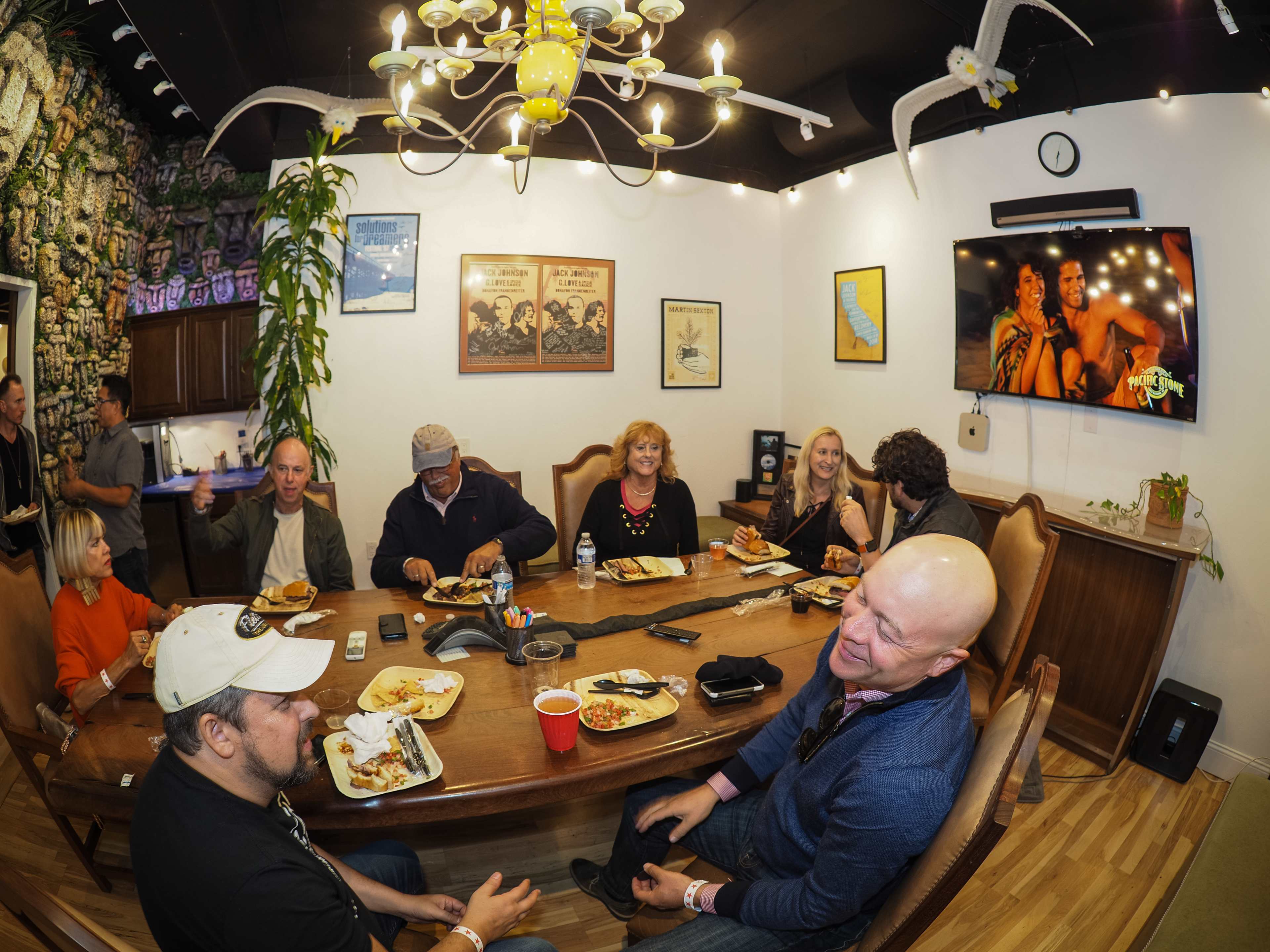 A group of people gathers around a large table, enjoying food and drinks while engaged in conversation in a brightly lit room decorated with various pictures and plants.
