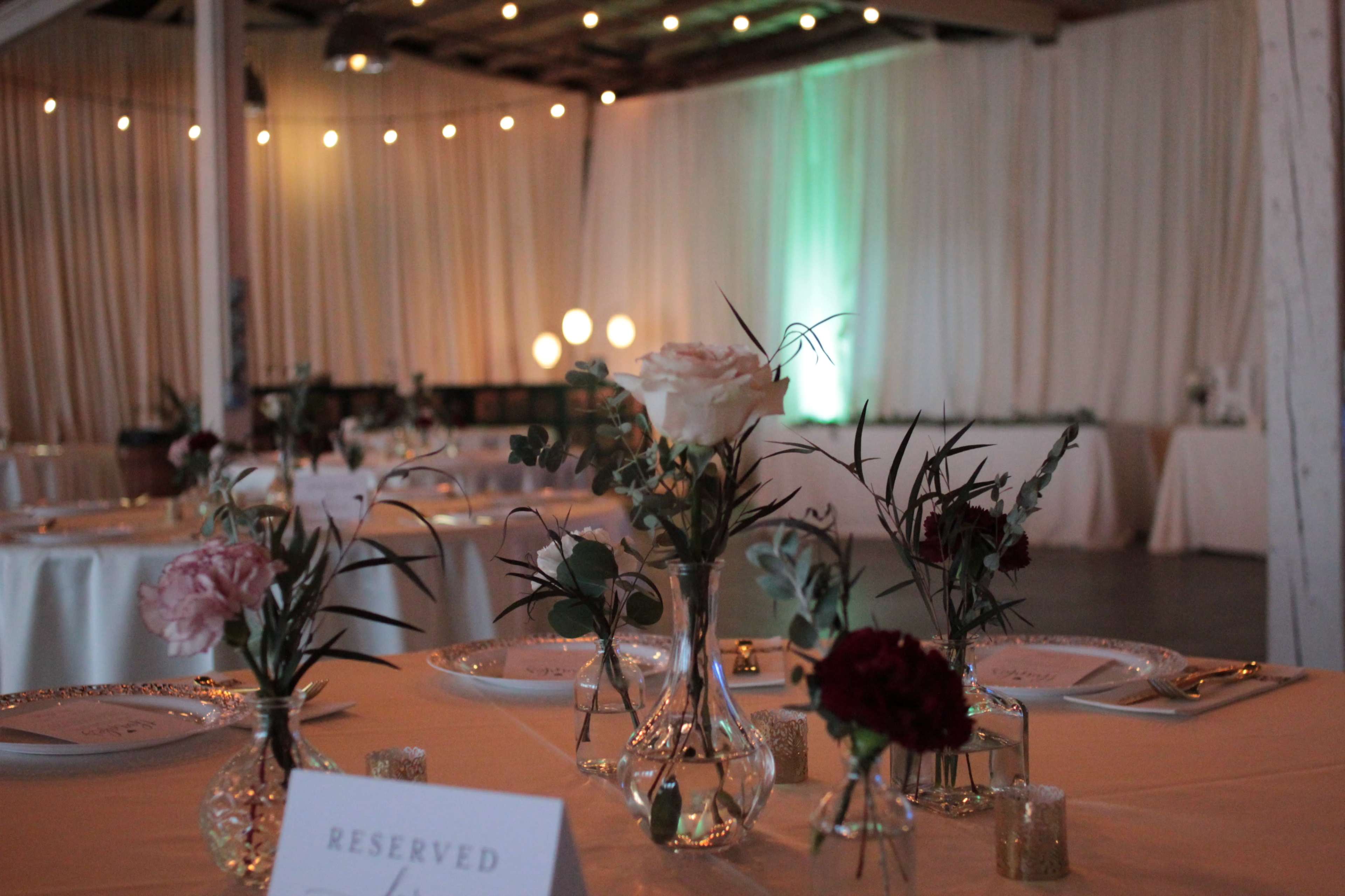 A beautifully set dining area with tables adorned with small floral arrangements and reserved signs, under draped fabric and string lights.