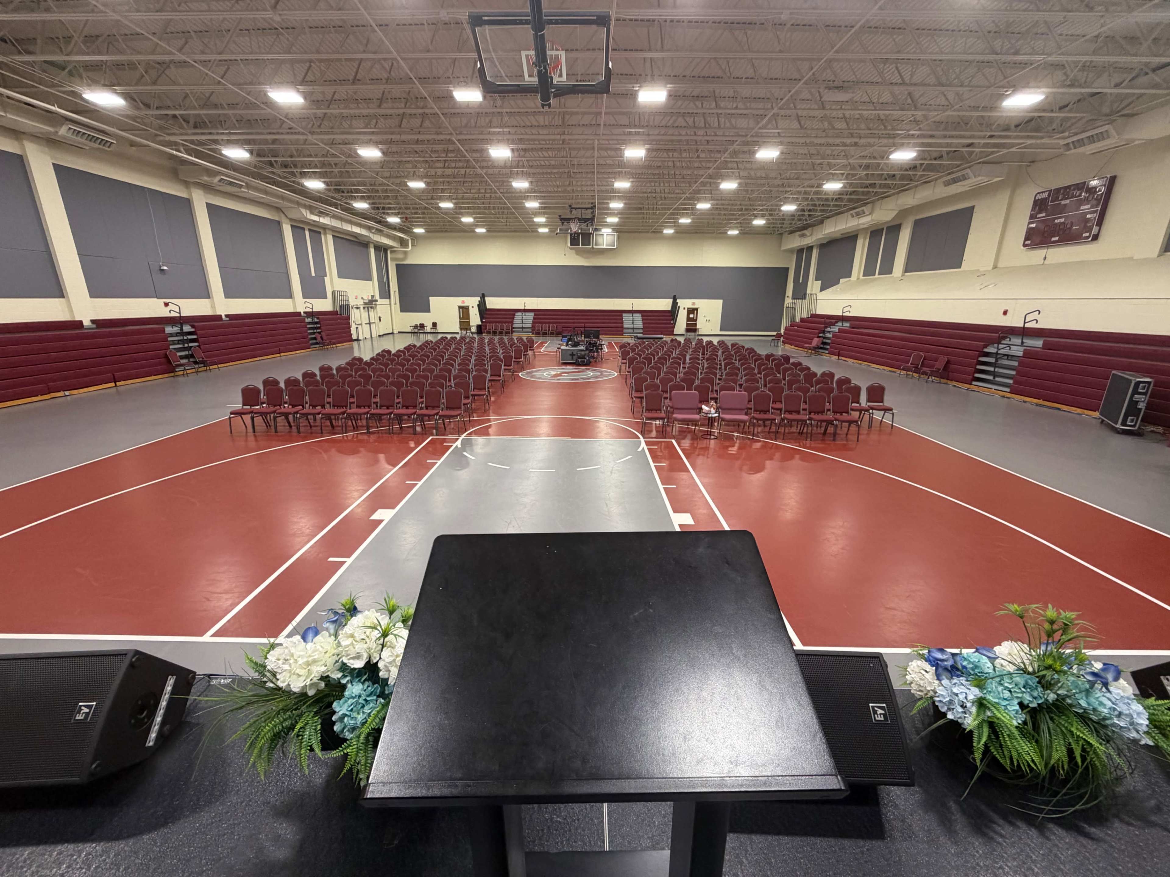 The image shows a gymnasium set up for an event, featuring rows of maroon chairs arranged in front of a podium on a basketball court.