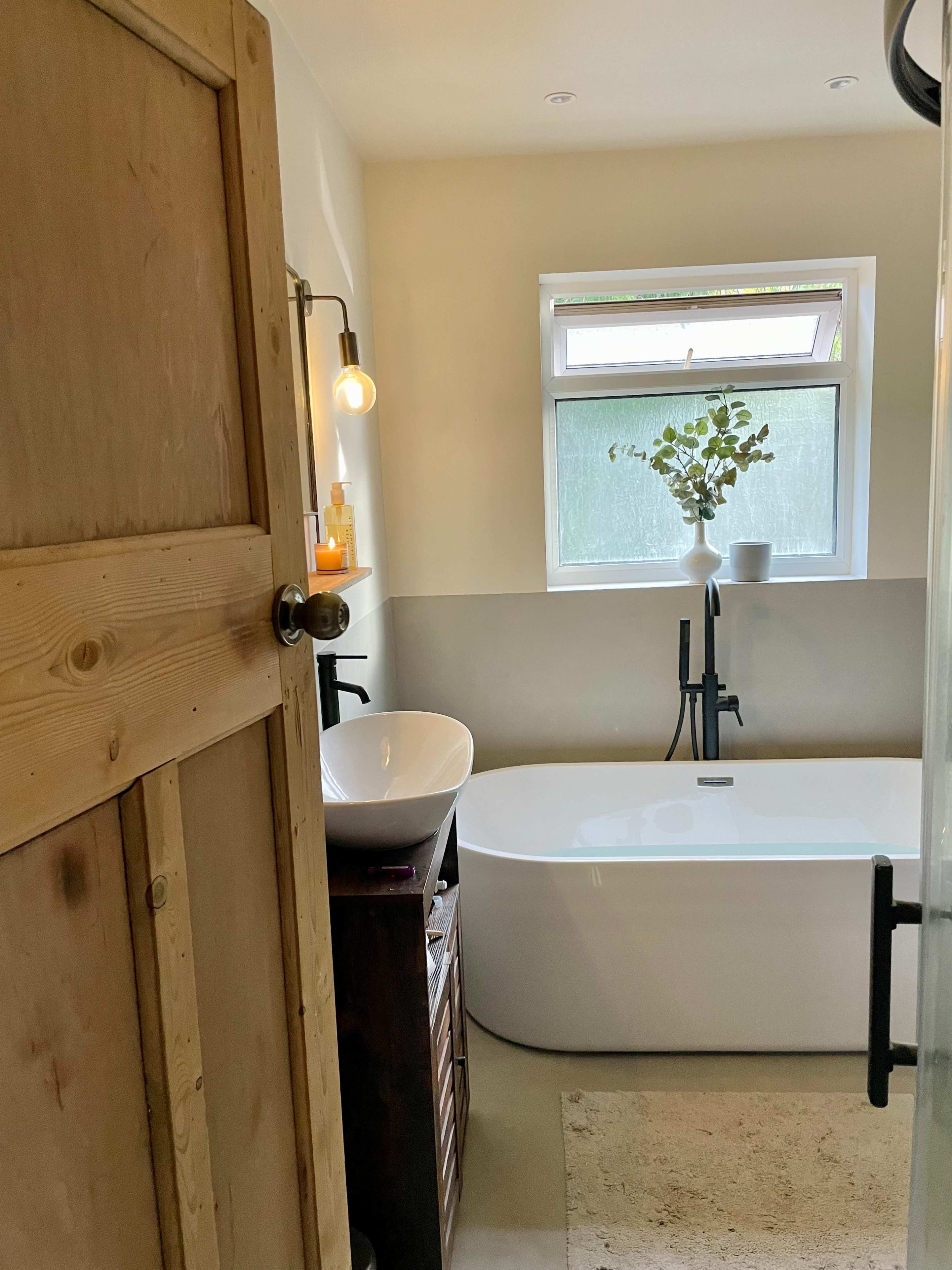 A modern bathroom featuring a freestanding bathtub, a sink with a wall-mounted faucet, and a window allowing natural light, along with a wooden door and minimal decor.
