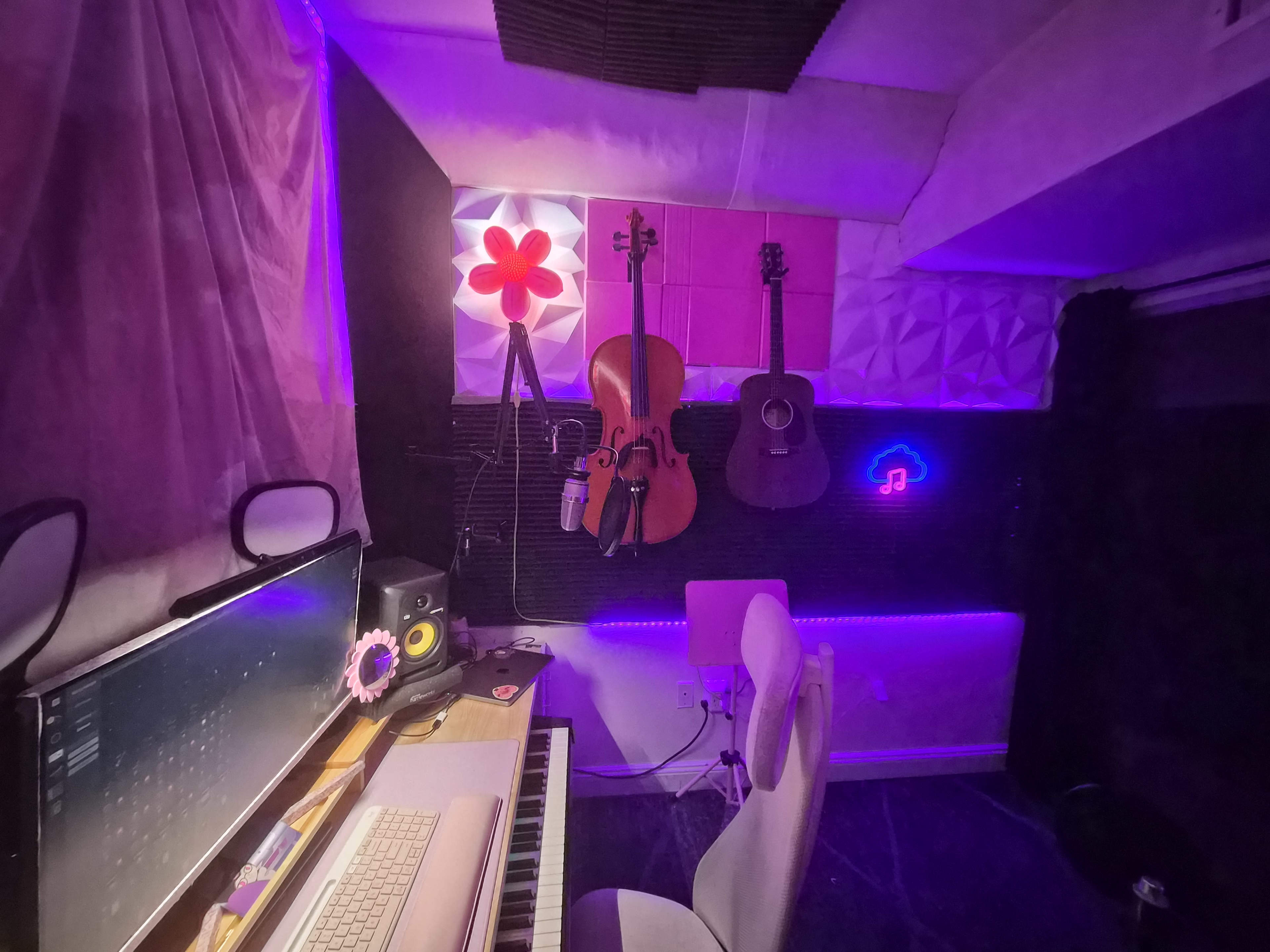 The image shows a small music studio featuring a computer setup with a keyboard, several musical instruments hanging on the wall, and LED lighting creating an ambient atmosphere.