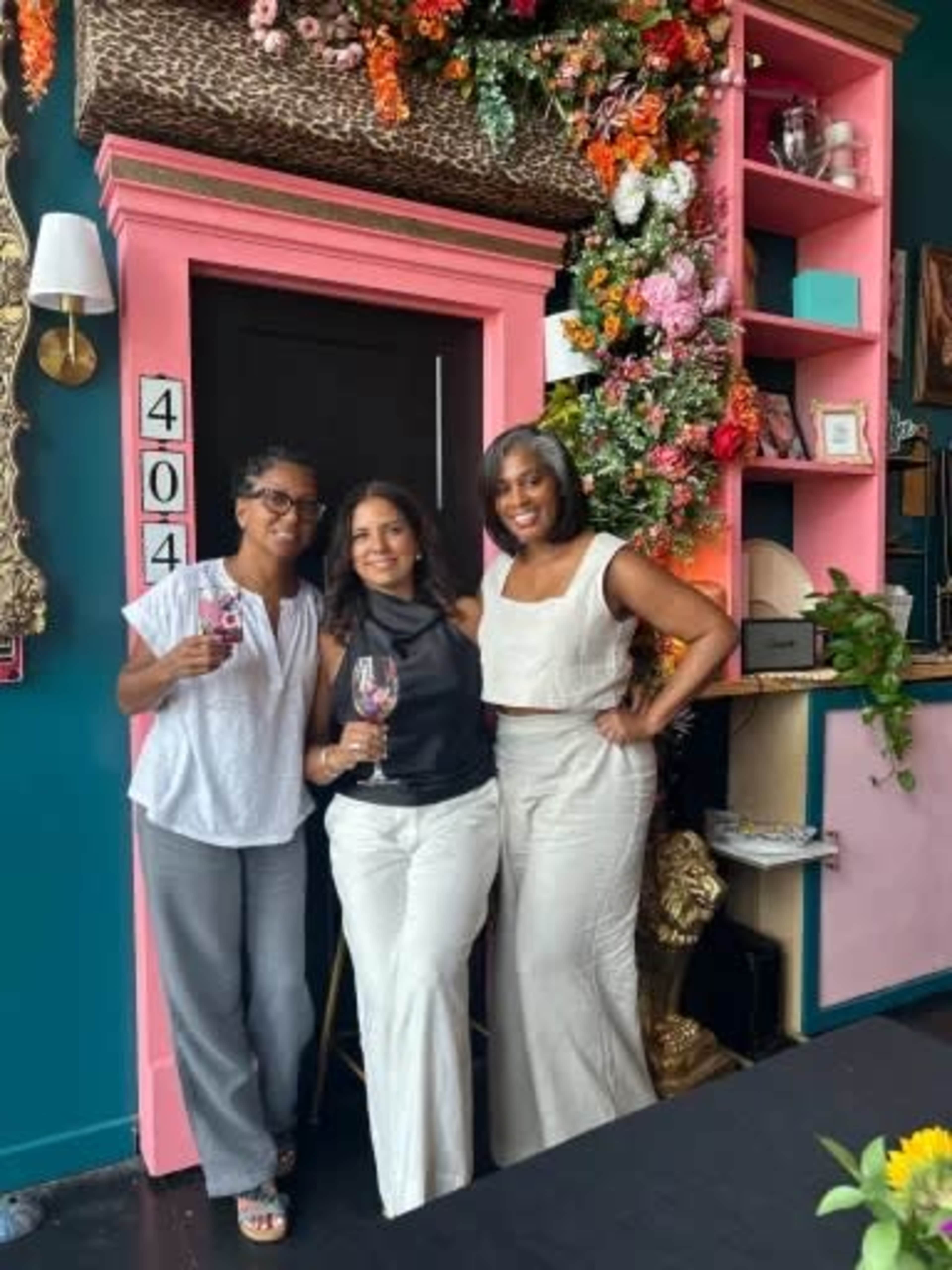 Three women stand together in a floral-decorated area, each holding a drink, in front of a vibrant pink and teal backdrop.