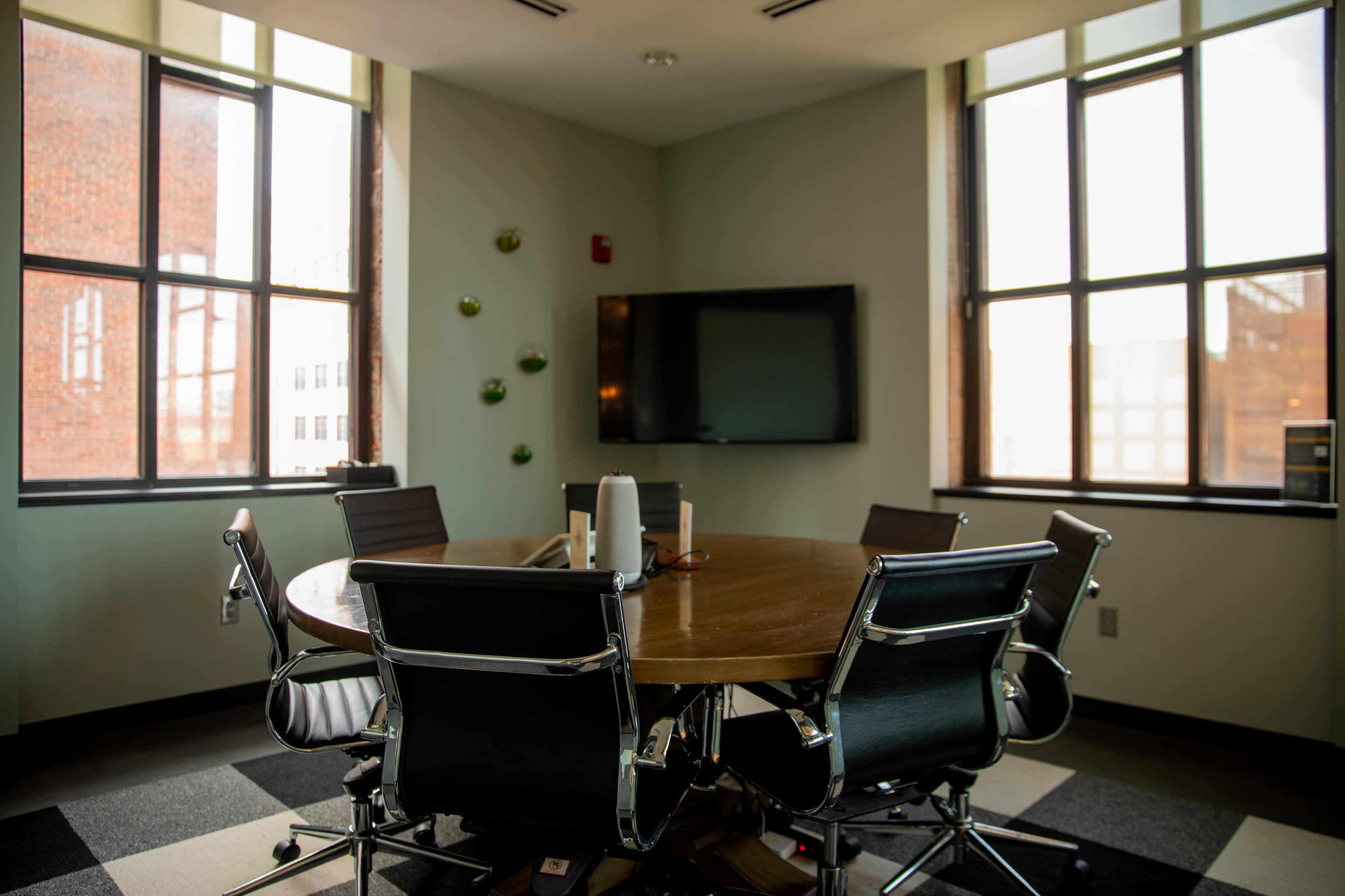 The image shows a round conference table surrounded by black leather chairs in a well-lit meeting room, featuring large windows and a wall-mounted television.