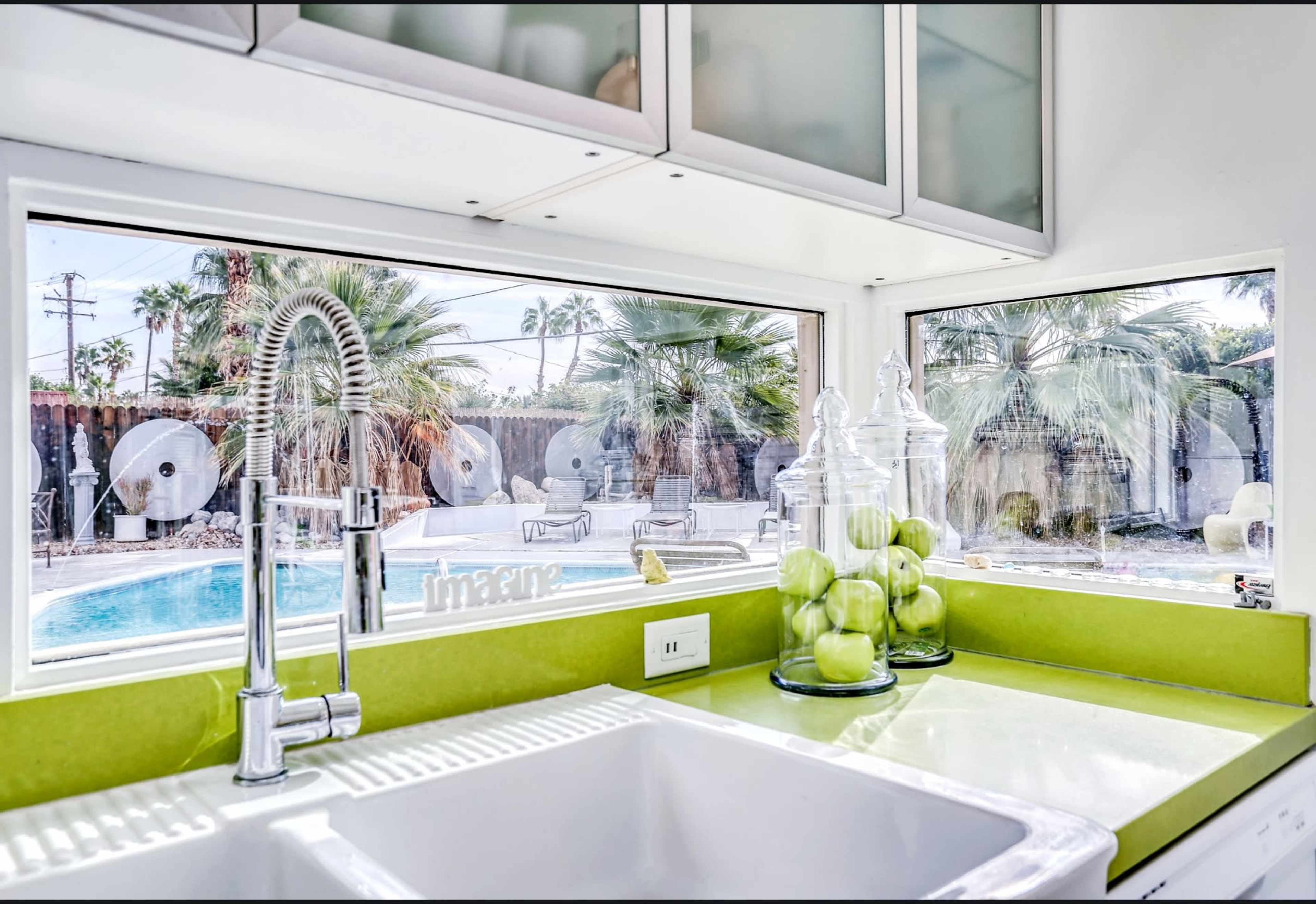 A bright kitchen features a green countertop, a white sink, and glass jars filled with green apples, with a view of a swimming pool and palm trees outside.