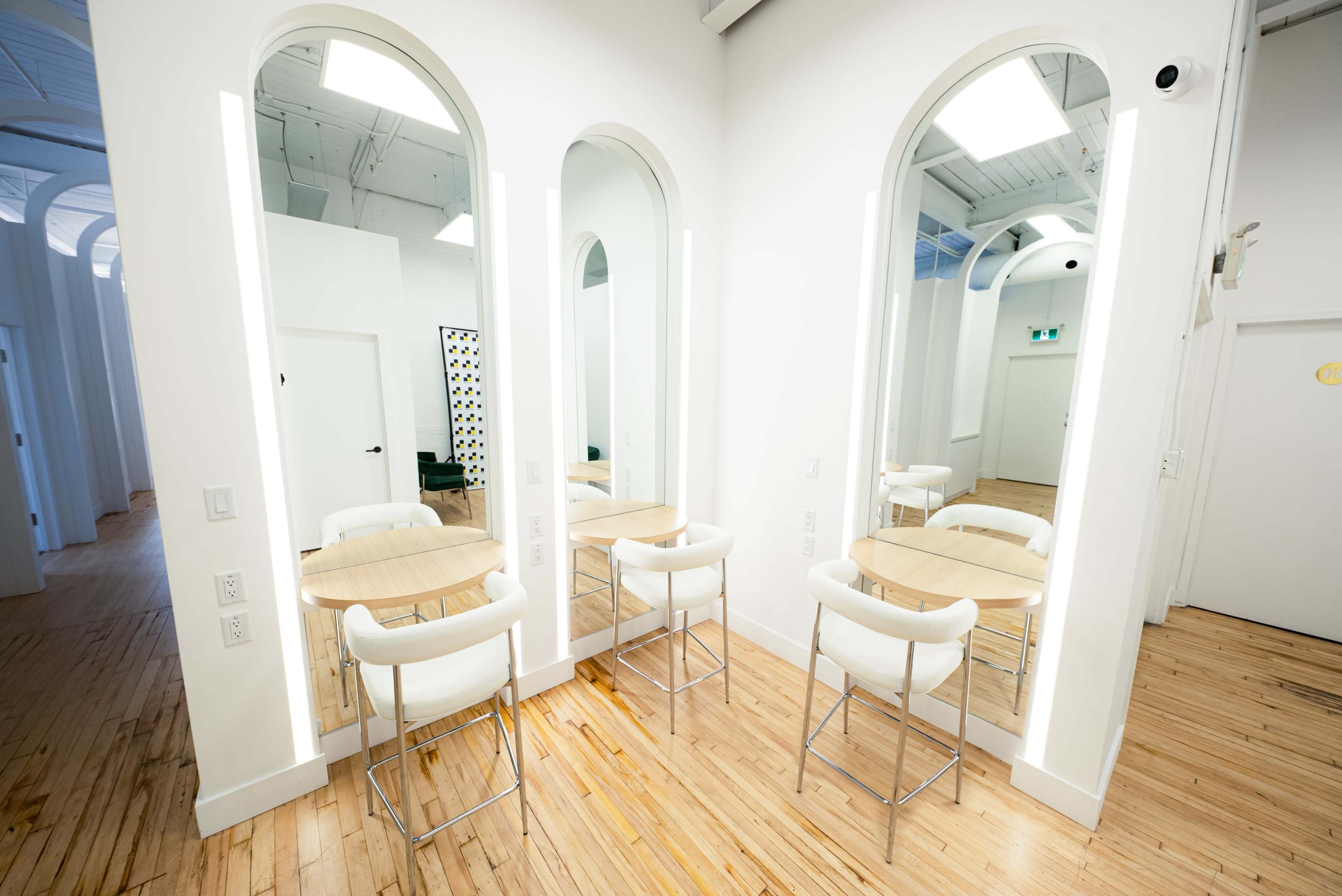 A bright, minimalist interior with three arch-shaped mirrors and two small round tables surrounded by white chairs on a wooden floor.