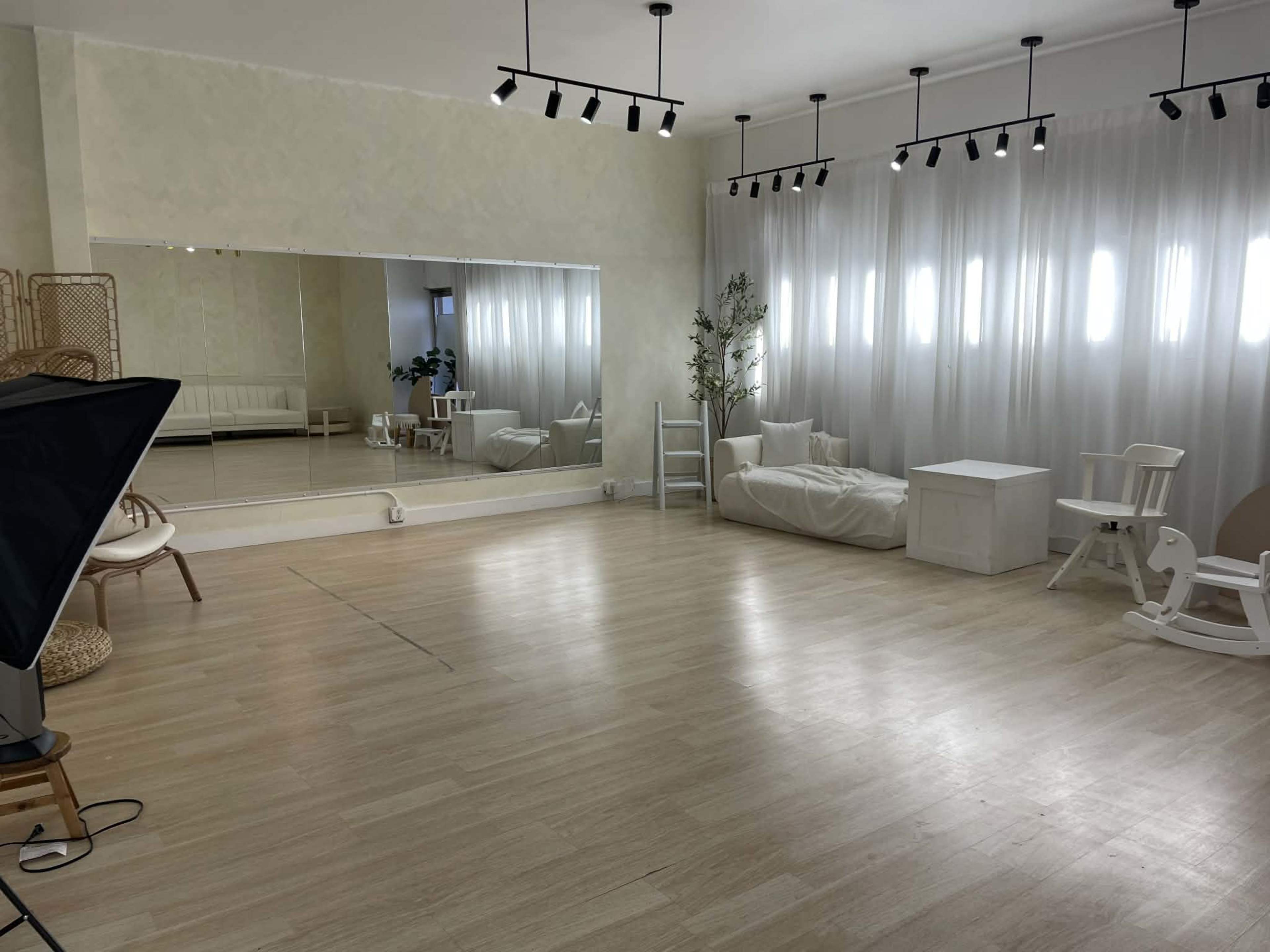 The image shows a spacious, well-lit room with wooden flooring, a large mirror on one wall, and white curtains allowing light to filter in.
