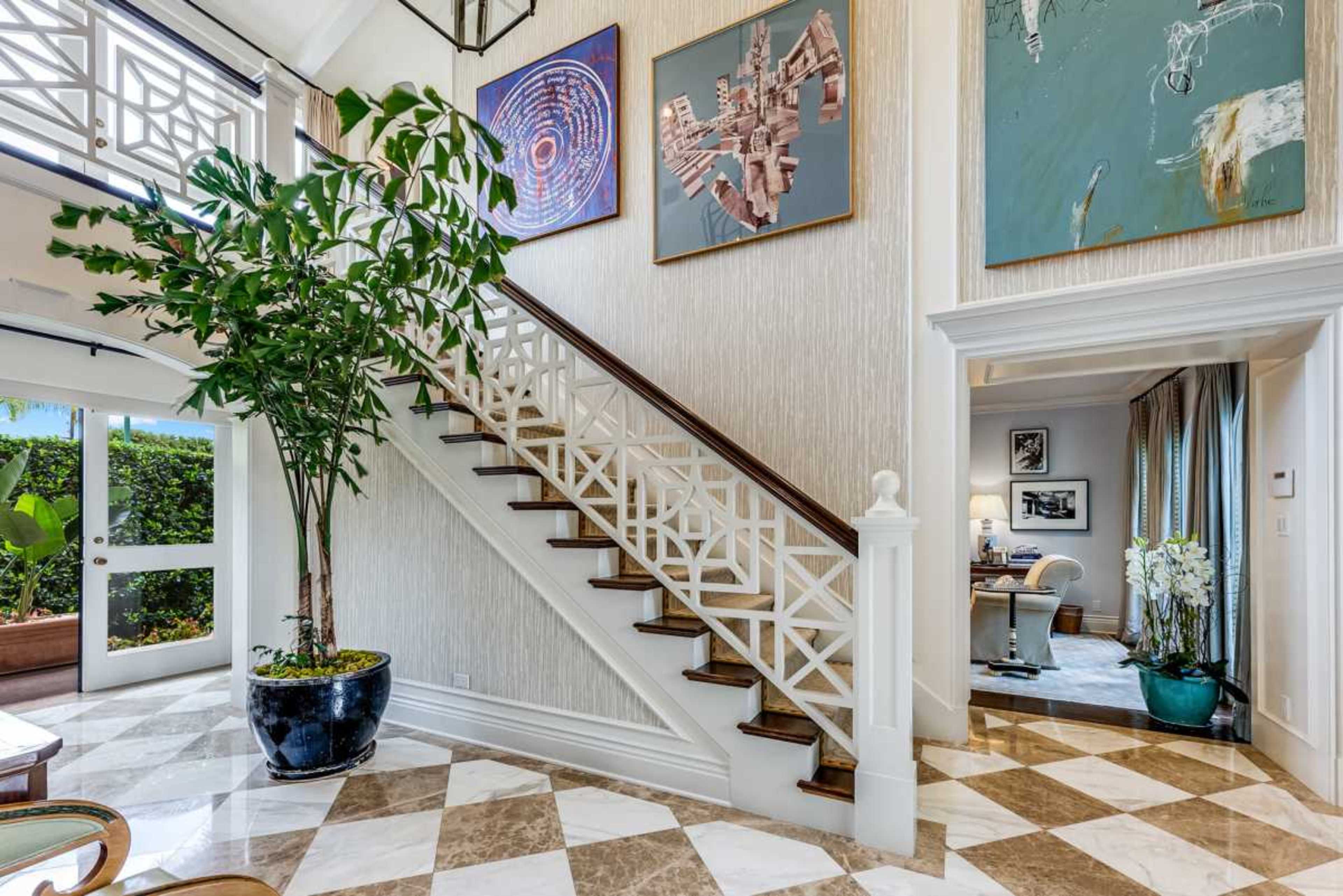 The image shows a spacious entryway with a staircase, decorated walls featuring artwork, and a large potted plant.
