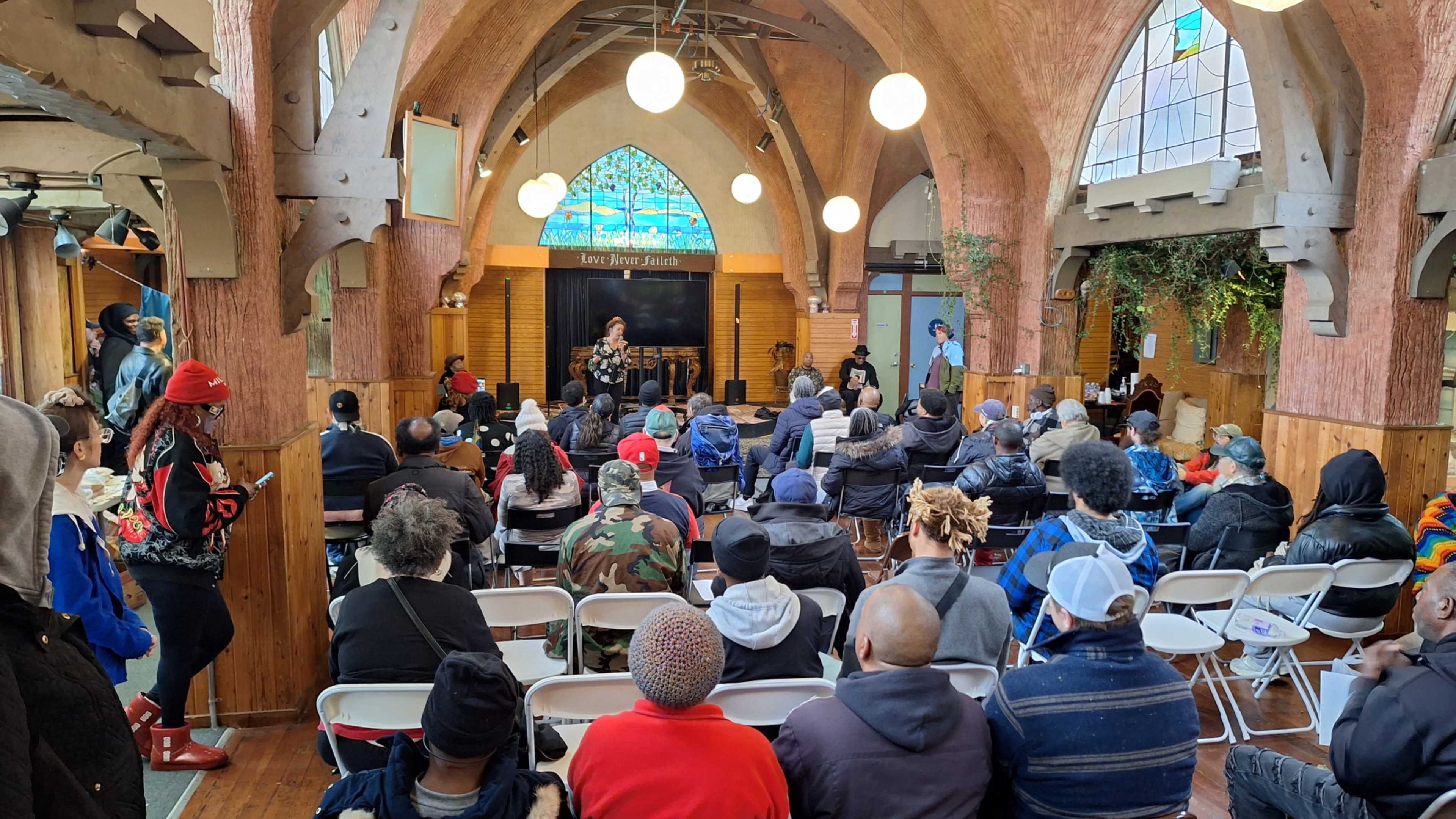 A large audience sits in a venue with arched wooden ceilings and colorful stained glass, attentively listening to speakers on a stage.