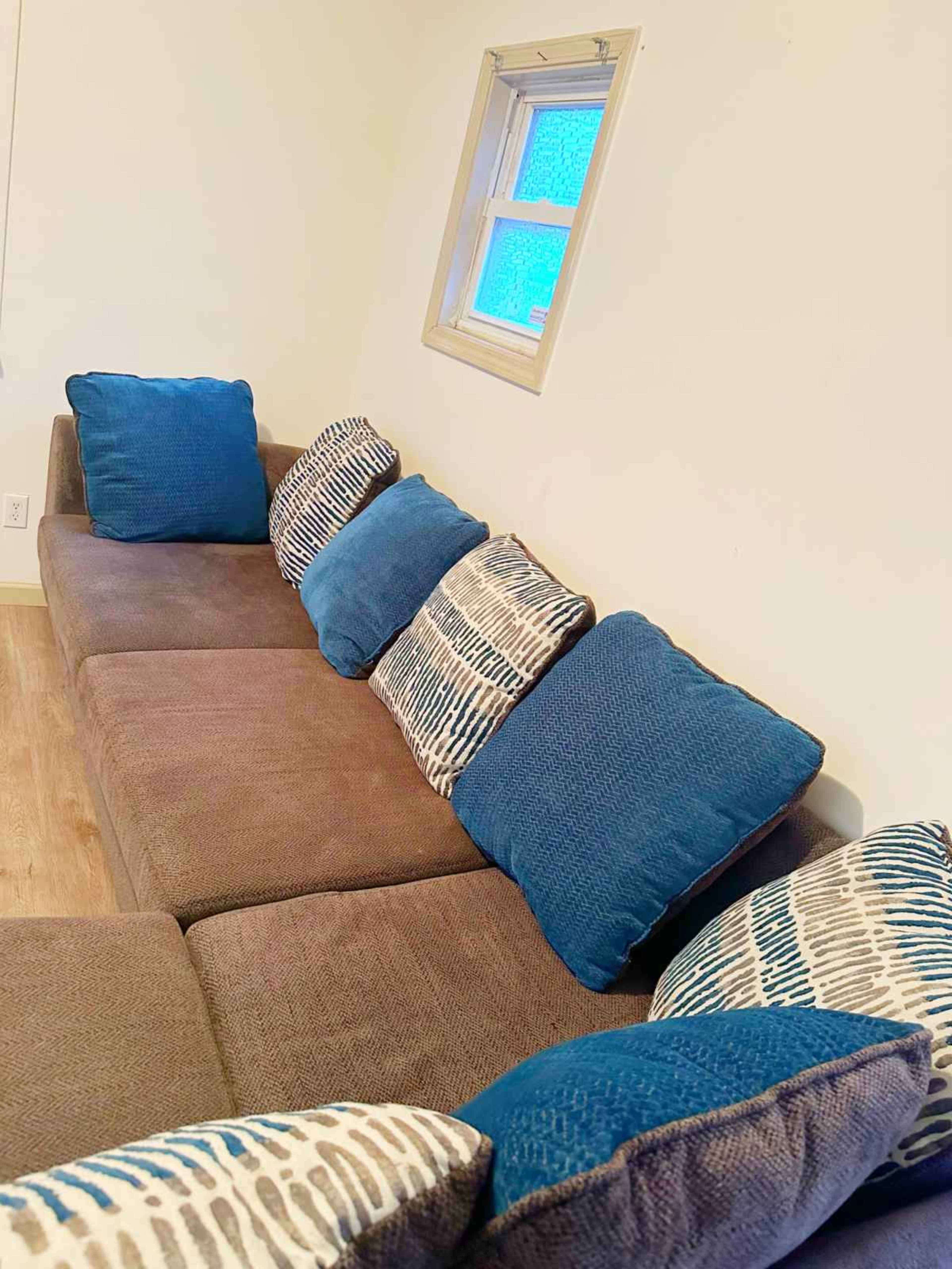 A brown couch with blue and patterned pillows is positioned against a light-colored wall beneath a small window.