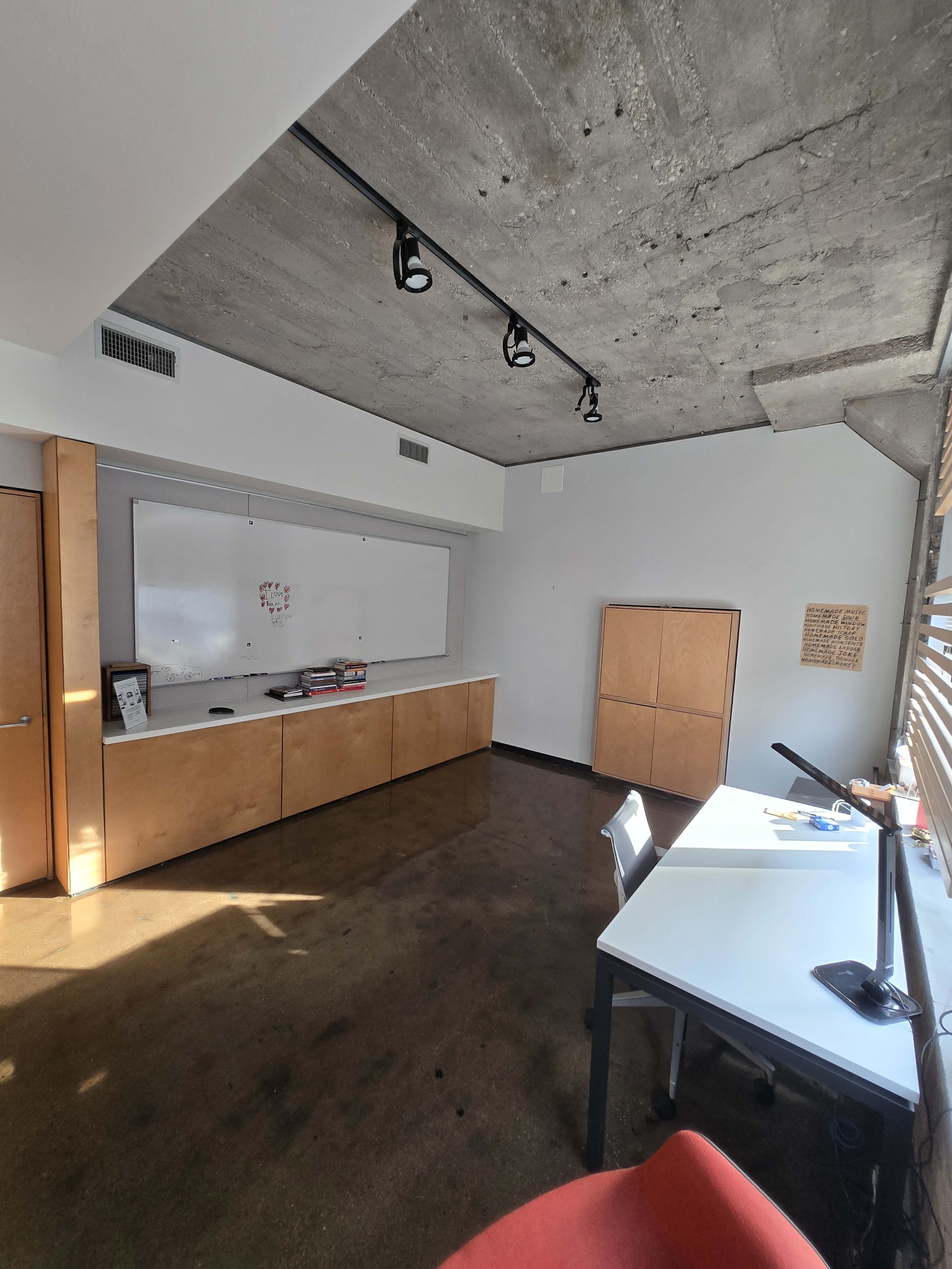 The image shows a modern office space with a concrete ceiling, a whiteboard on the wall, a desk with a computer, and storage cabinets.