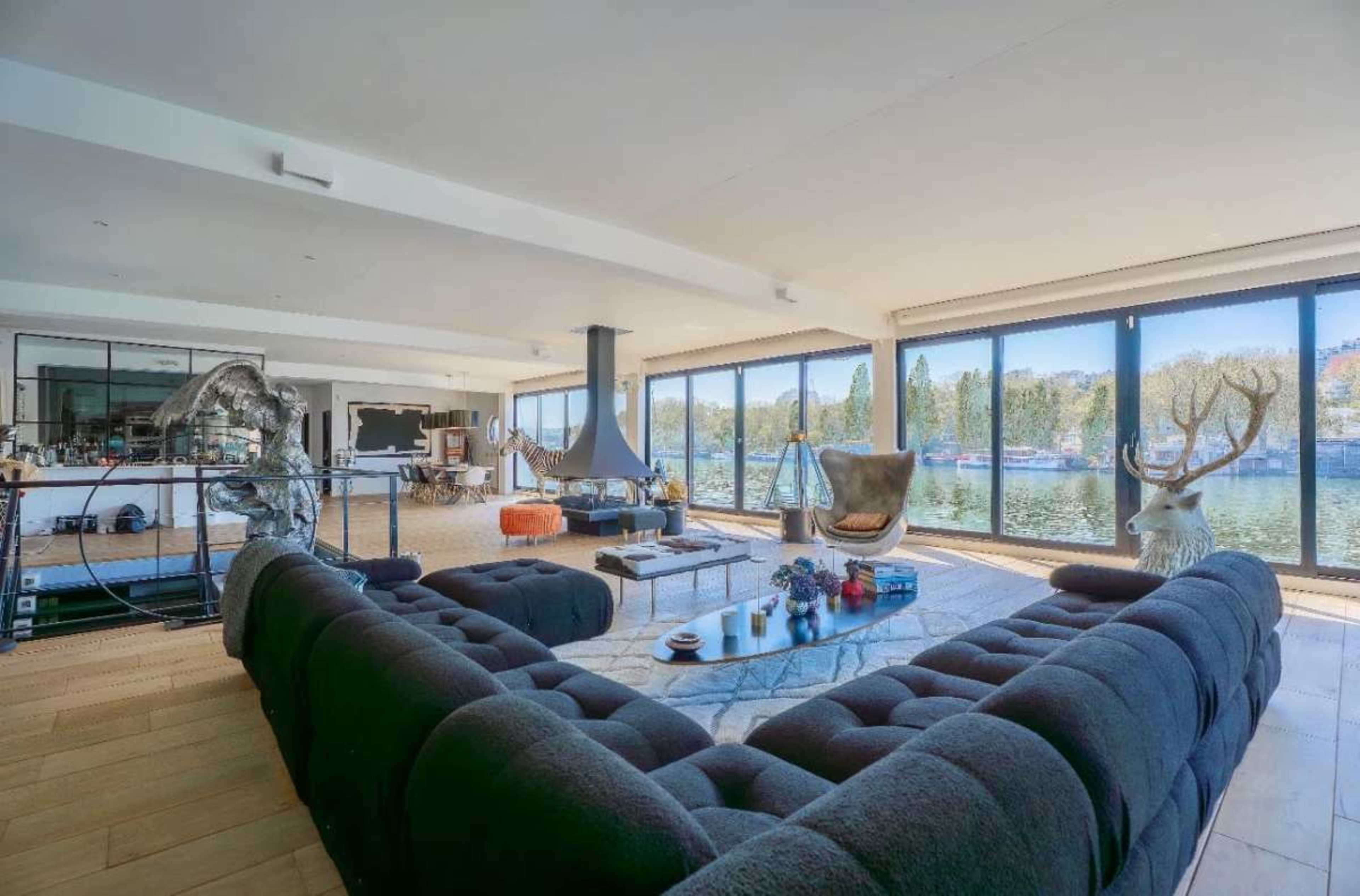The image shows a modern living room with a large dark sofa facing a wall of windows that overlook a body of water.