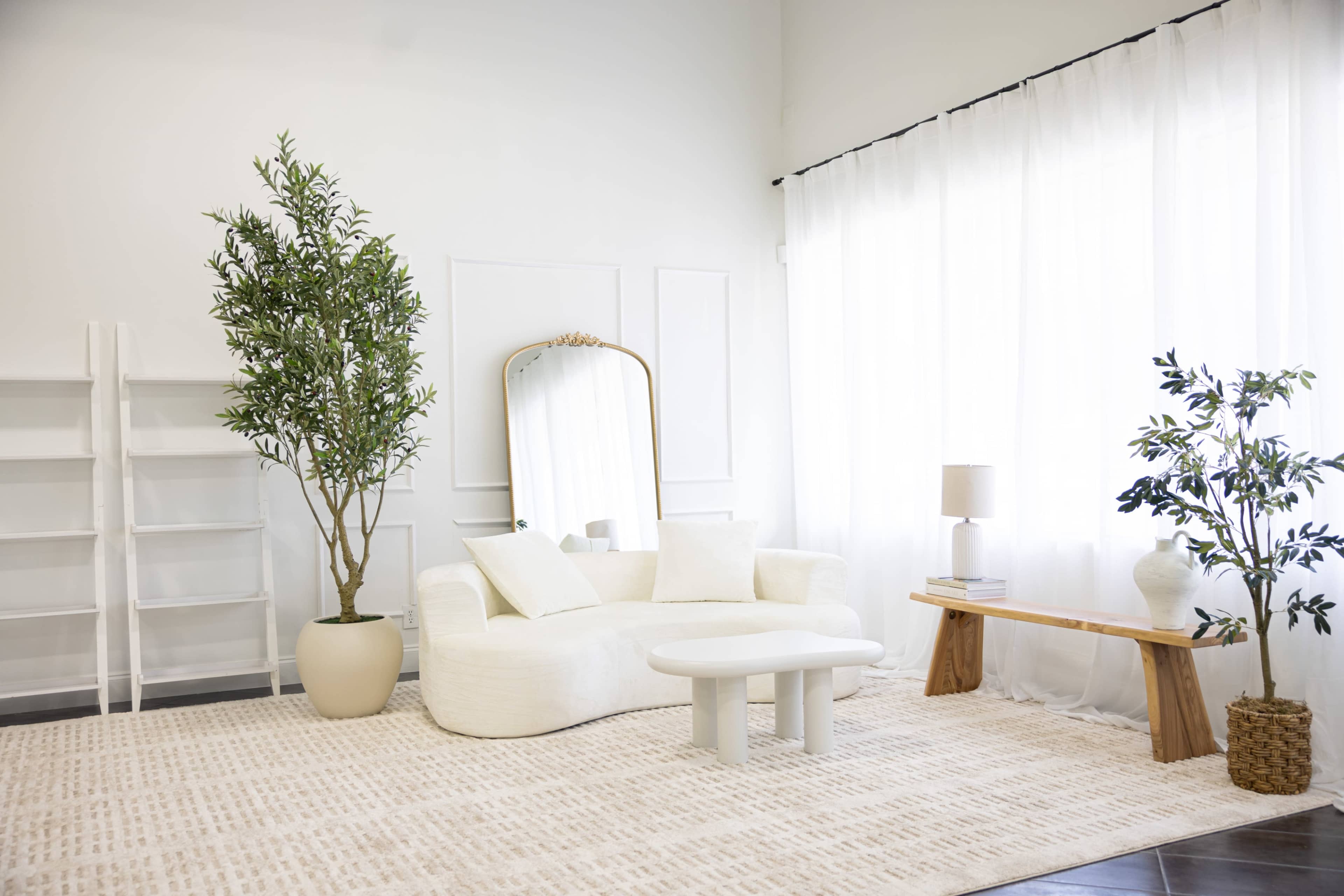 The image depicts a modern living room with a white sofa, a round coffee table, a wooden side table, potted plants, and large windows covered with sheer curtains.