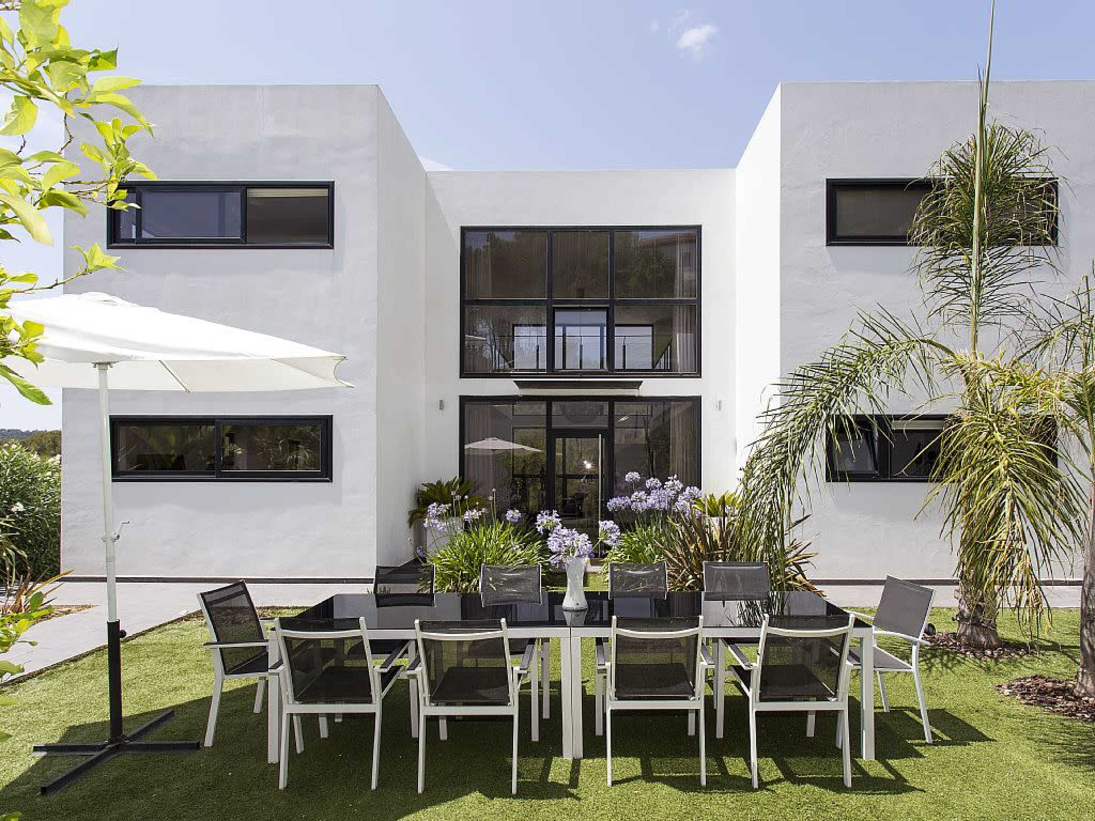A modern two-story house with large windows features a spacious outdoor dining area set with a long table and chairs on a grassy lawn.