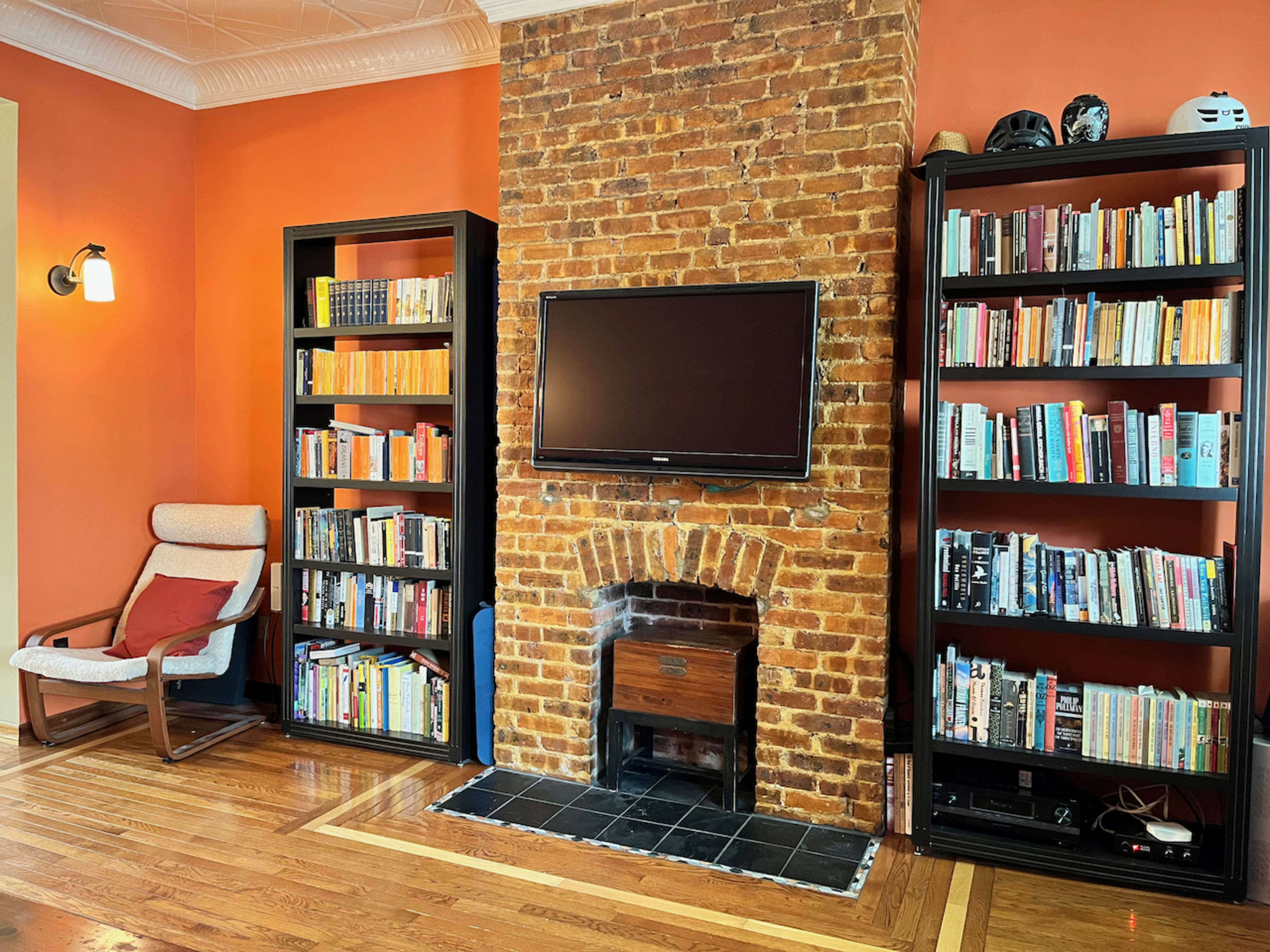 A living room features a brick fireplace with a television mounted above it, flanked by two bookshelves filled with books, against a bright orange wall.